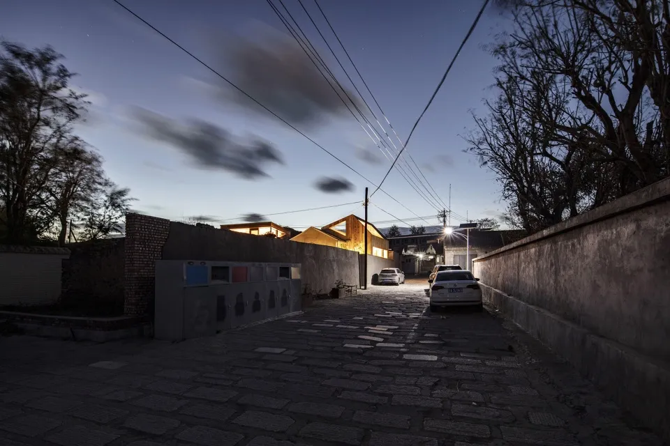 A cobblestone street at dusk with buildings and cars illuminated by lights.