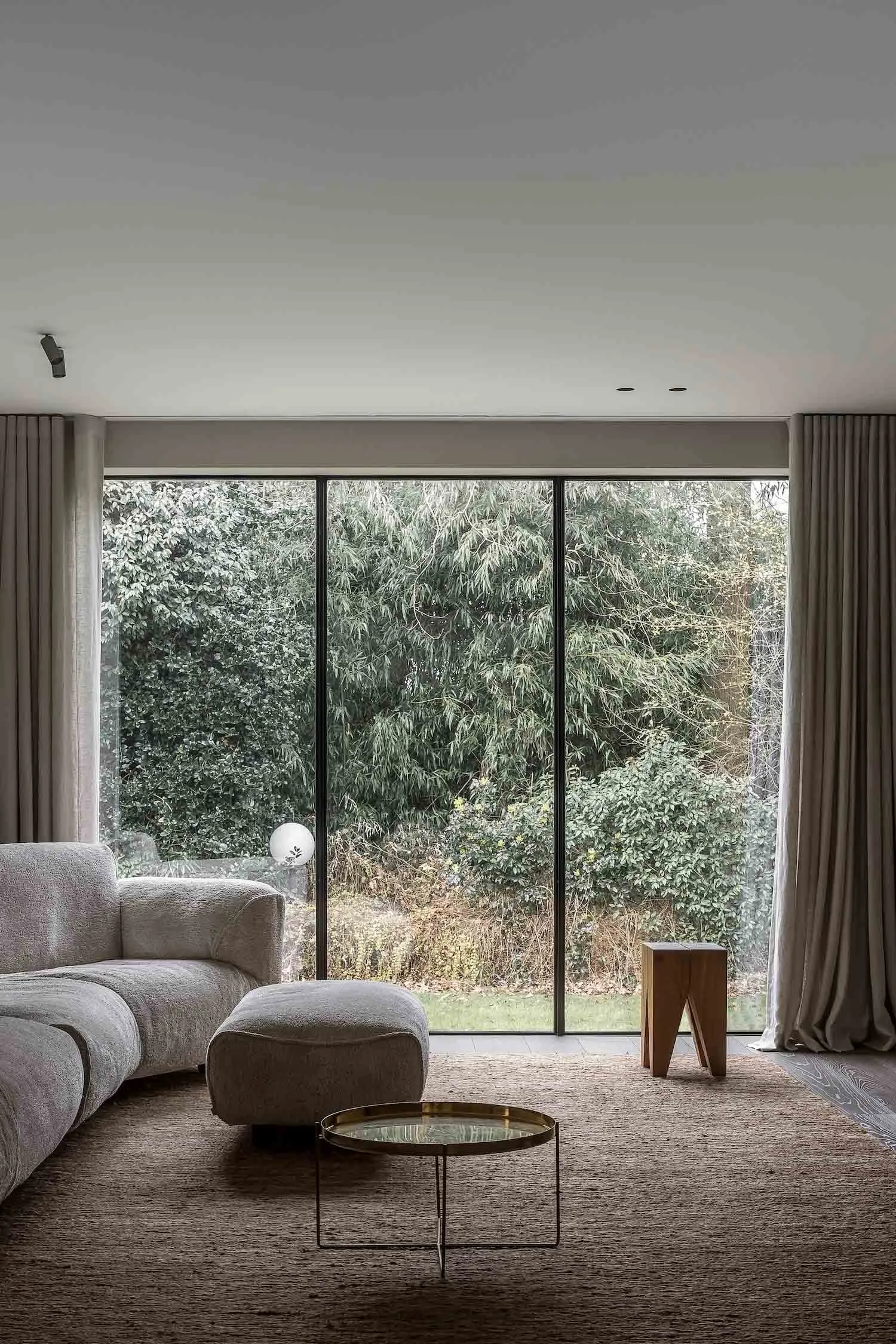 A minimalist living room with a gray fabric sofa, a low coffee table, and a wood stool, featuring large windows with views of
