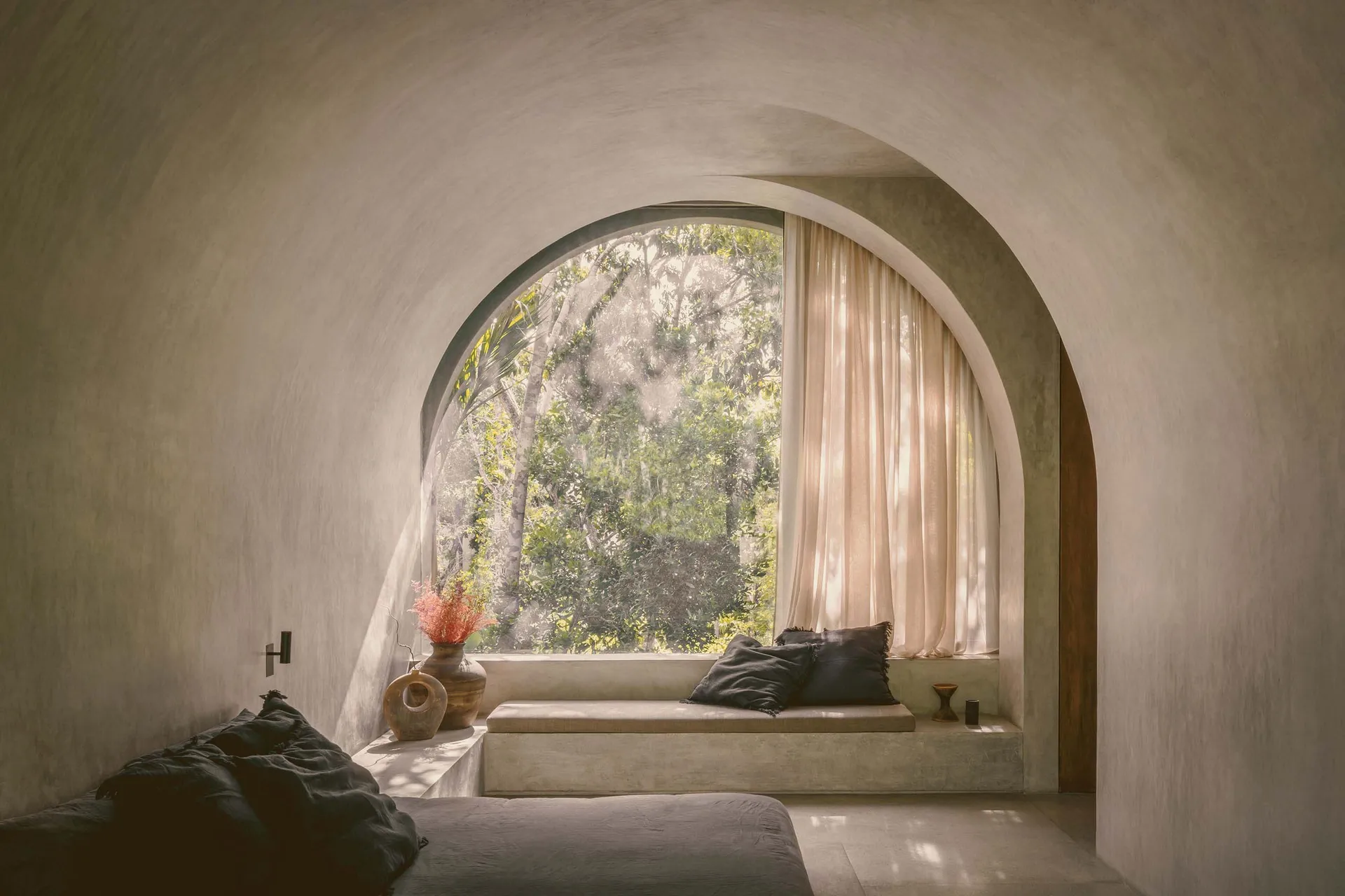 Modern bedroom featuring a concrete sitting area with natural light from a large arched window overlooking trees.