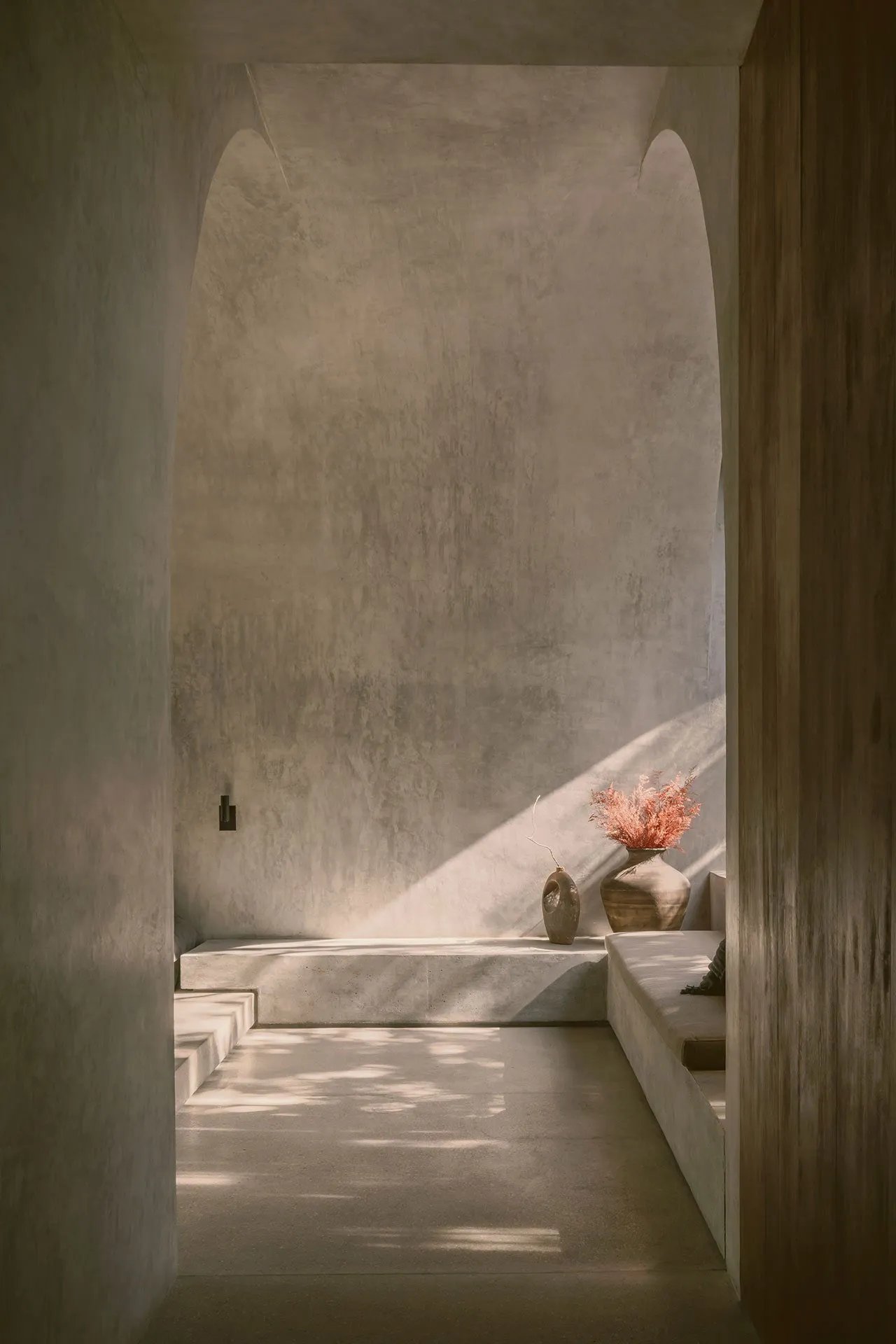 Interior of a minimalist space with concrete walls and floor, featuring a vase with dried flowers and a bench.