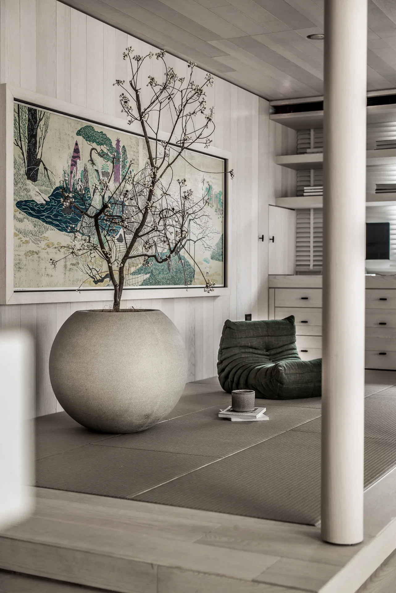 A serene living room with wooden floors, a large decorative pot with plants, and a minimalist green seat.
