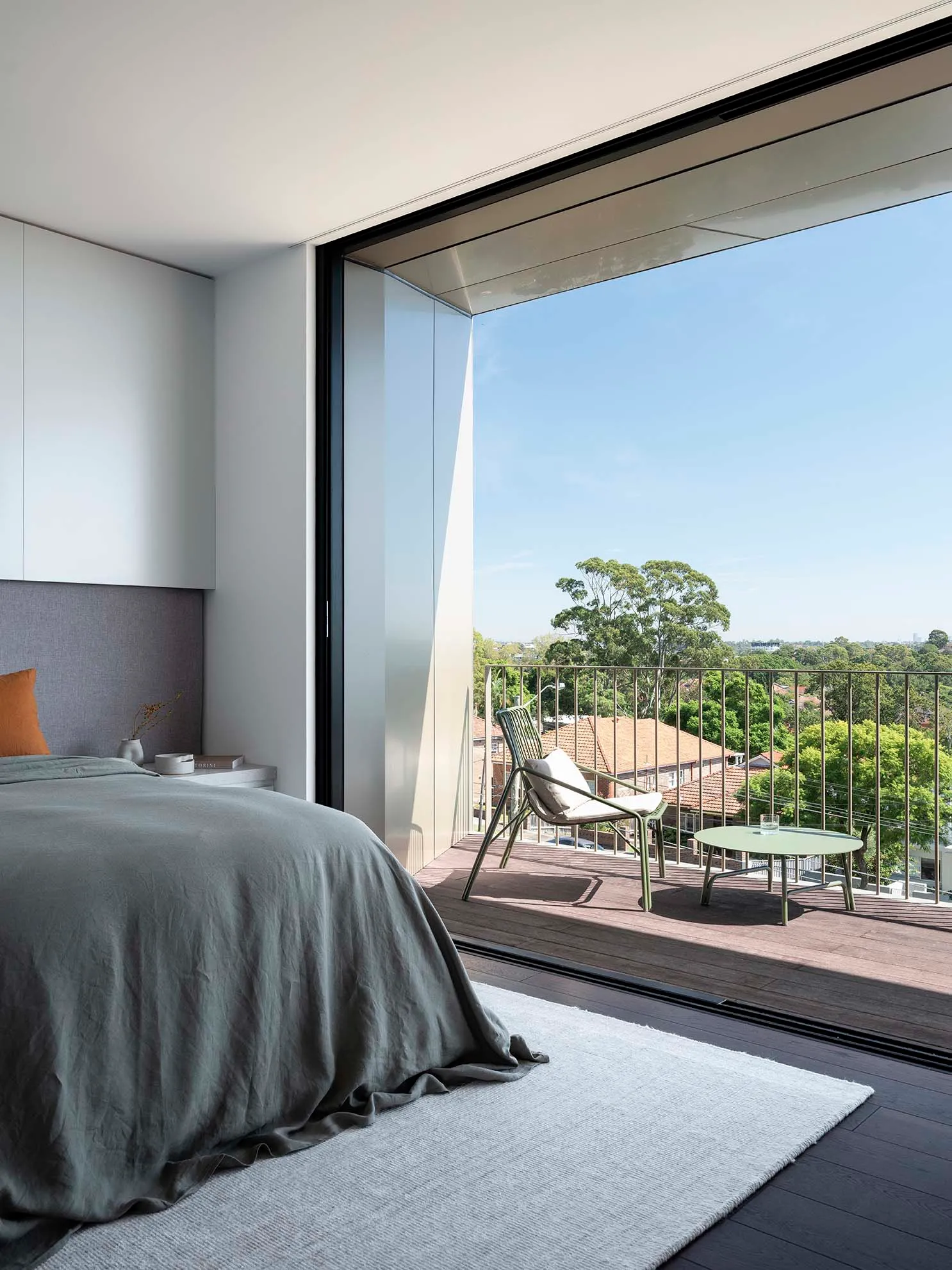 A modern bedroom with a wooden floor, featuring a bed, a rug, and a large window overlooking a balcony with a chair and table
