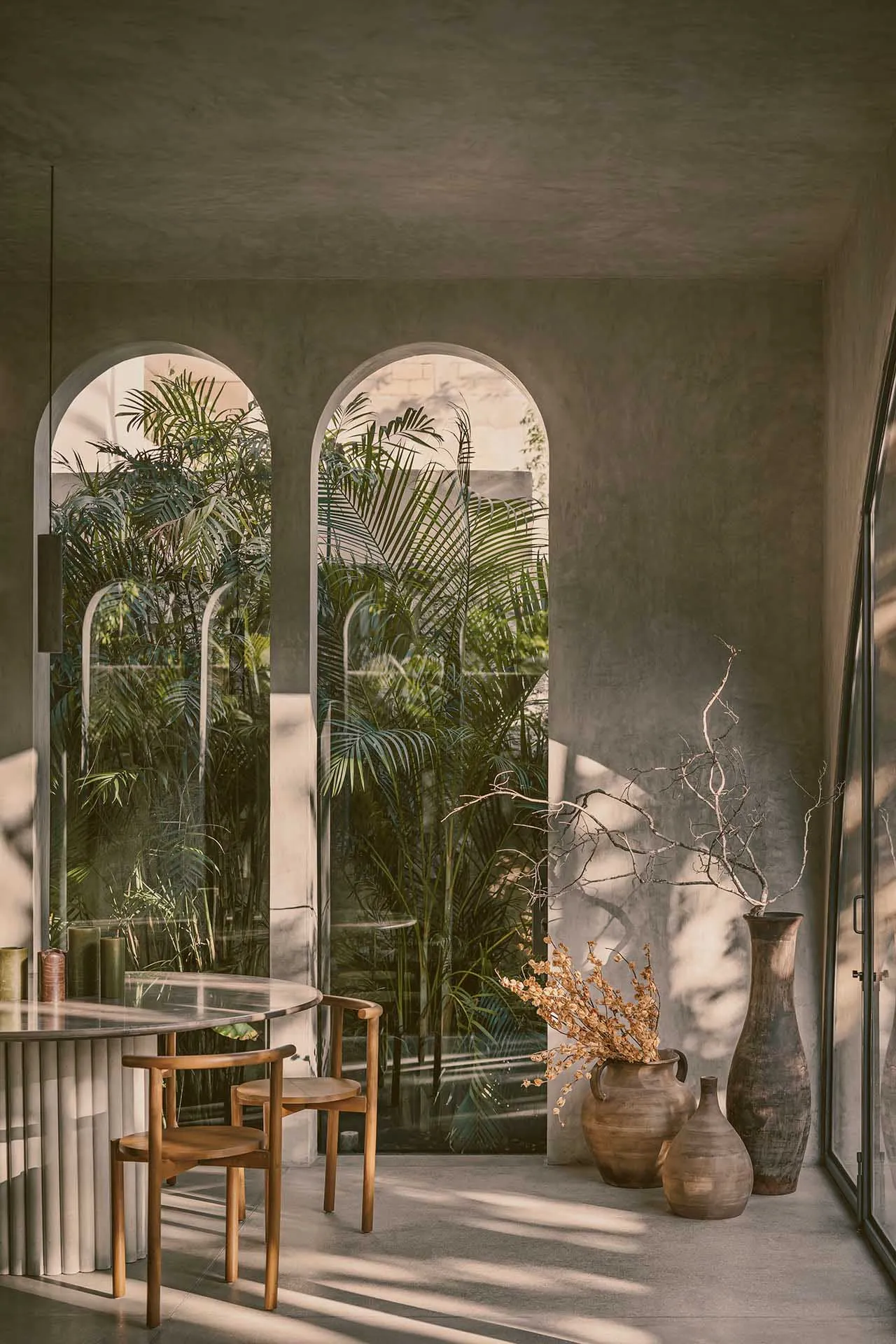 A modern, minimalist living room with concrete walls and floor, featuring a clear view of palm trees outside through arched w