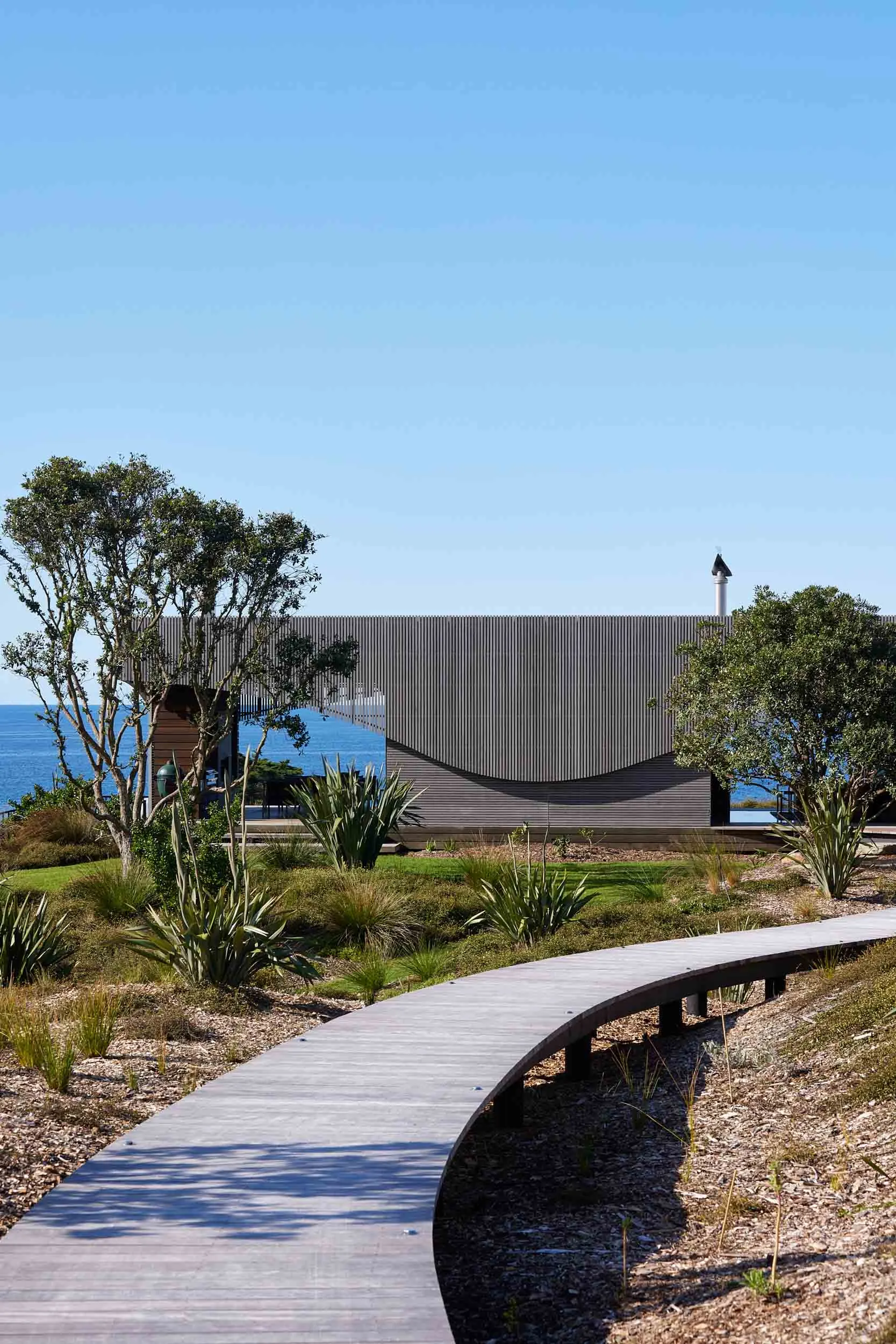 A modern architectural design featuring metal and wood materials, set against a clear blue sky with a serene ocean view.