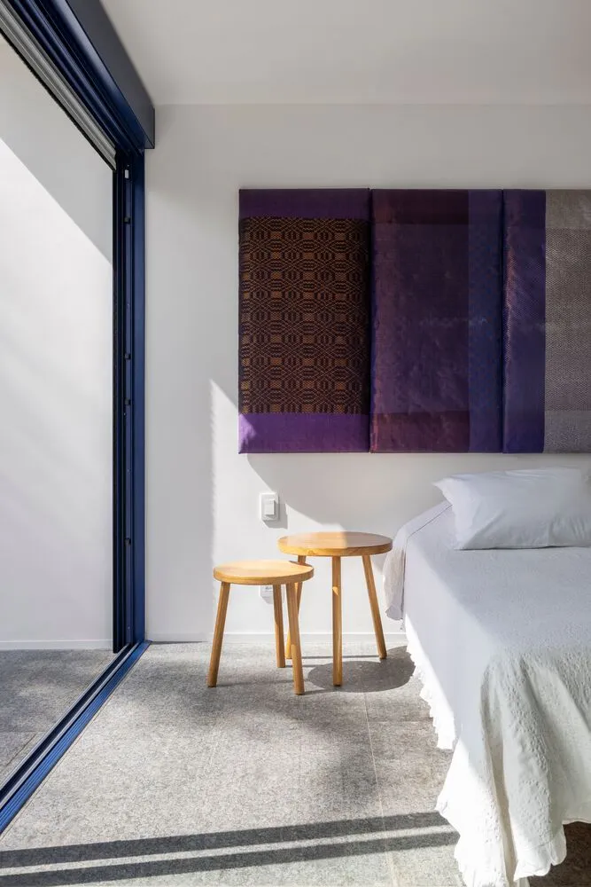 A modern bedroom featuring a bed with gray bedding, wooden stools, and a wall art piece with purple and brown panels.