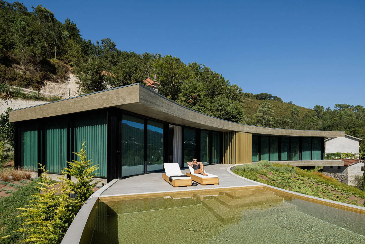 A modern holiday villa featuring a concrete and glass exterior with a swimming pool and lounge chairs.