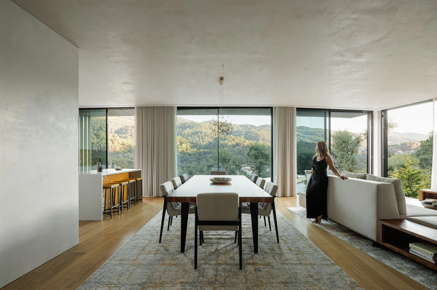 A modern minimalist dining room with a large table, chairs, and a view of the mountains through floor-to-ceiling windows.