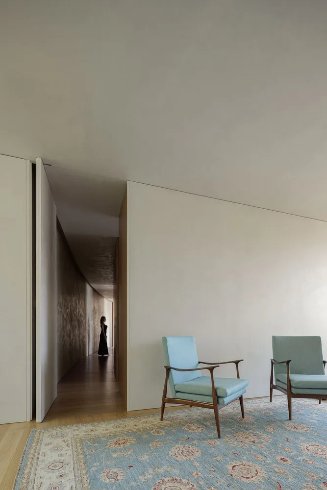 A minimalist living room with fabric-covered walls and wooden furniture, featuring a blue armchair on a floral-patterned rug.