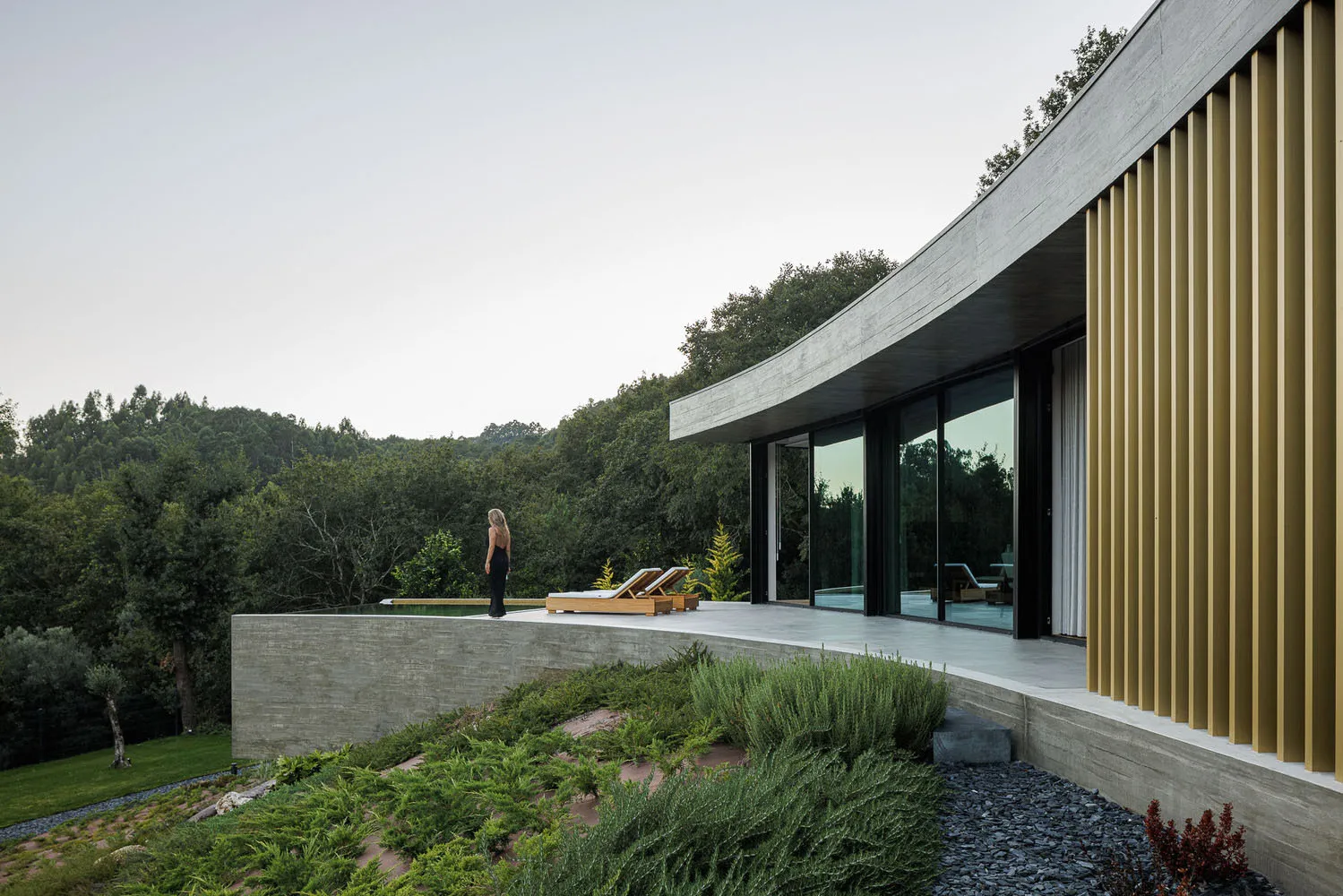 A modern residential exterior with concrete walls and large glass windows overlooking a forested area with two lounge chairs