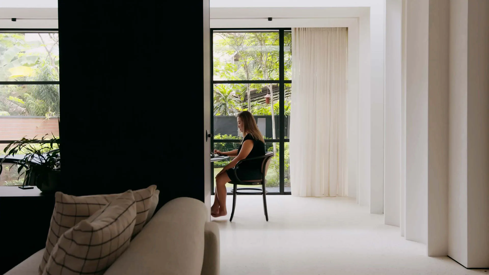 A modern home office with white walls, floor, and furniture, featuring a woman working on a laptop at a round table.