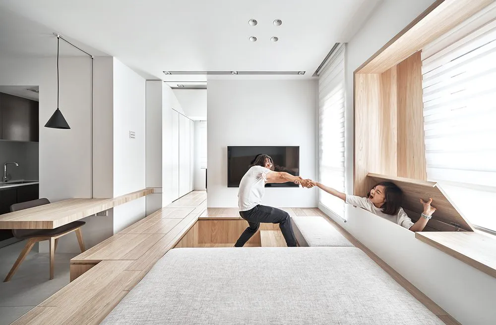 Two people interacting in a modern interior space with wooden benches and white walls, light wood flooring, and a window view