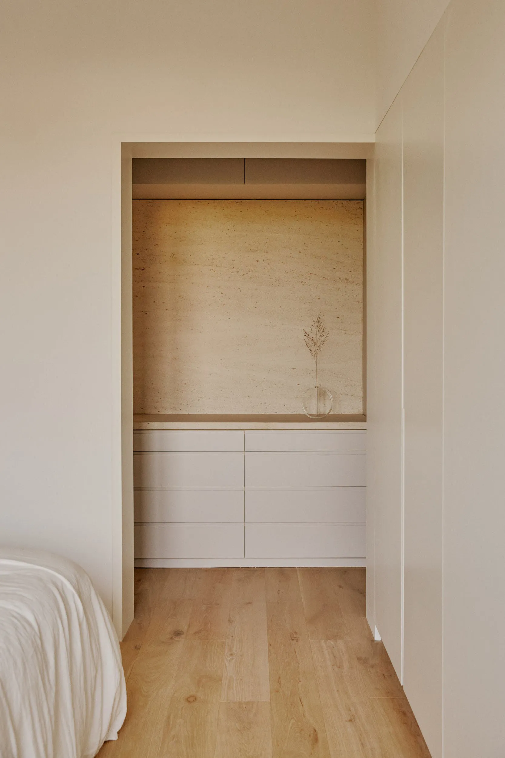 A minimalist bedroom storage unit featuring neutral-toned drawers and a vase with a dried plant against a natural wood floor 