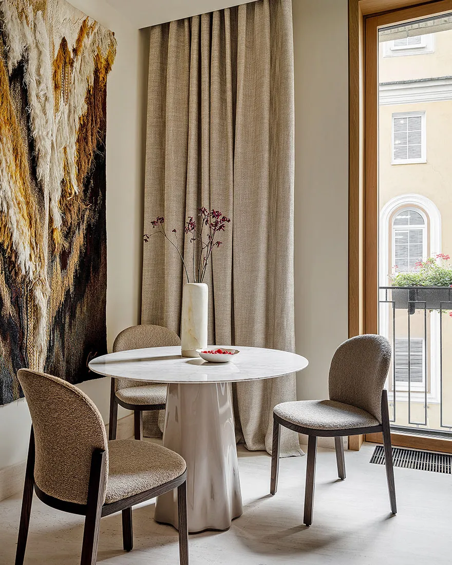 Modern dining room furnished with a round marble table and fabric chairs, featuring a decorative wall hanging and a vase with