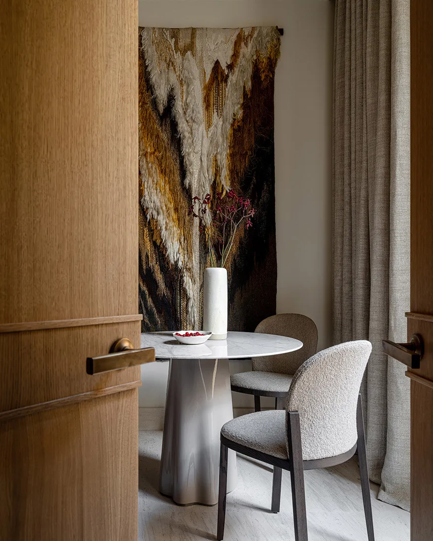 A modern dining room featuring a round marble table with white chairs, a vase with dried flowers, and a textured abstract art