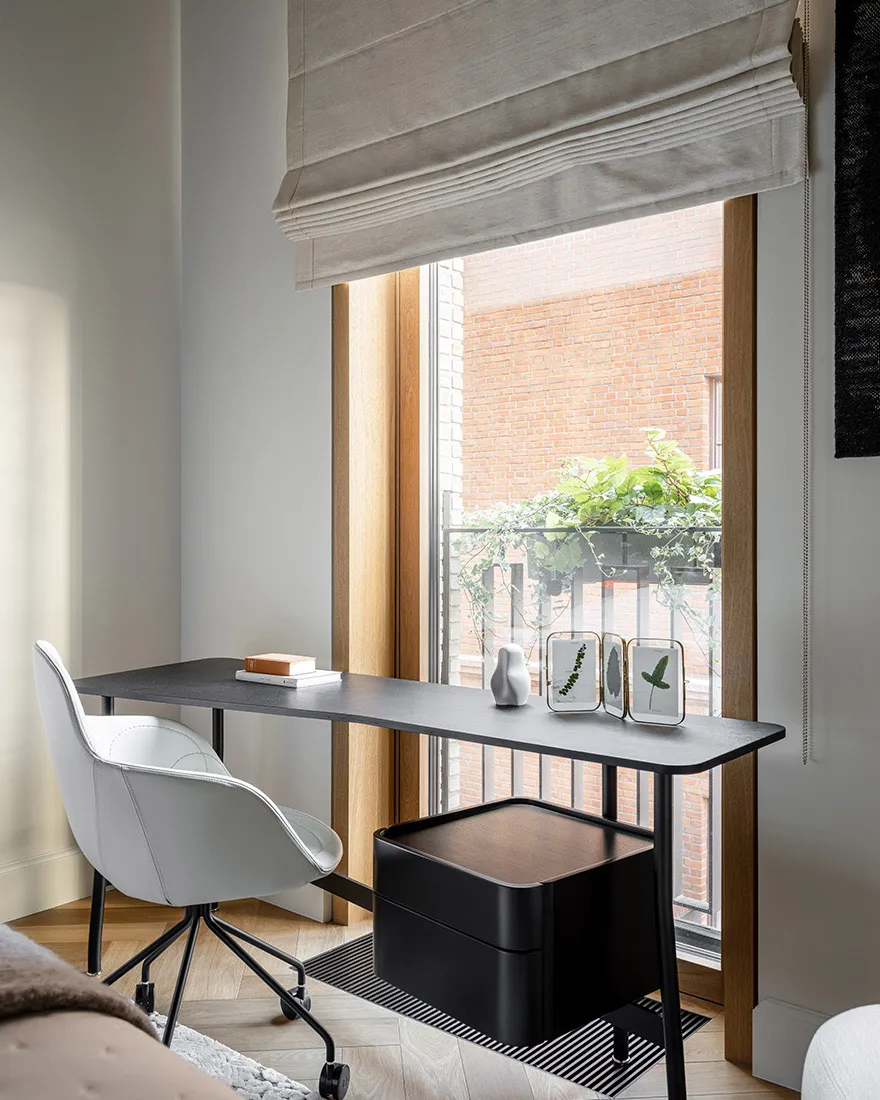 A minimalist home office featuring a sleek black desk, white ergonomic chair, and a window overlooking a red-brick building w
