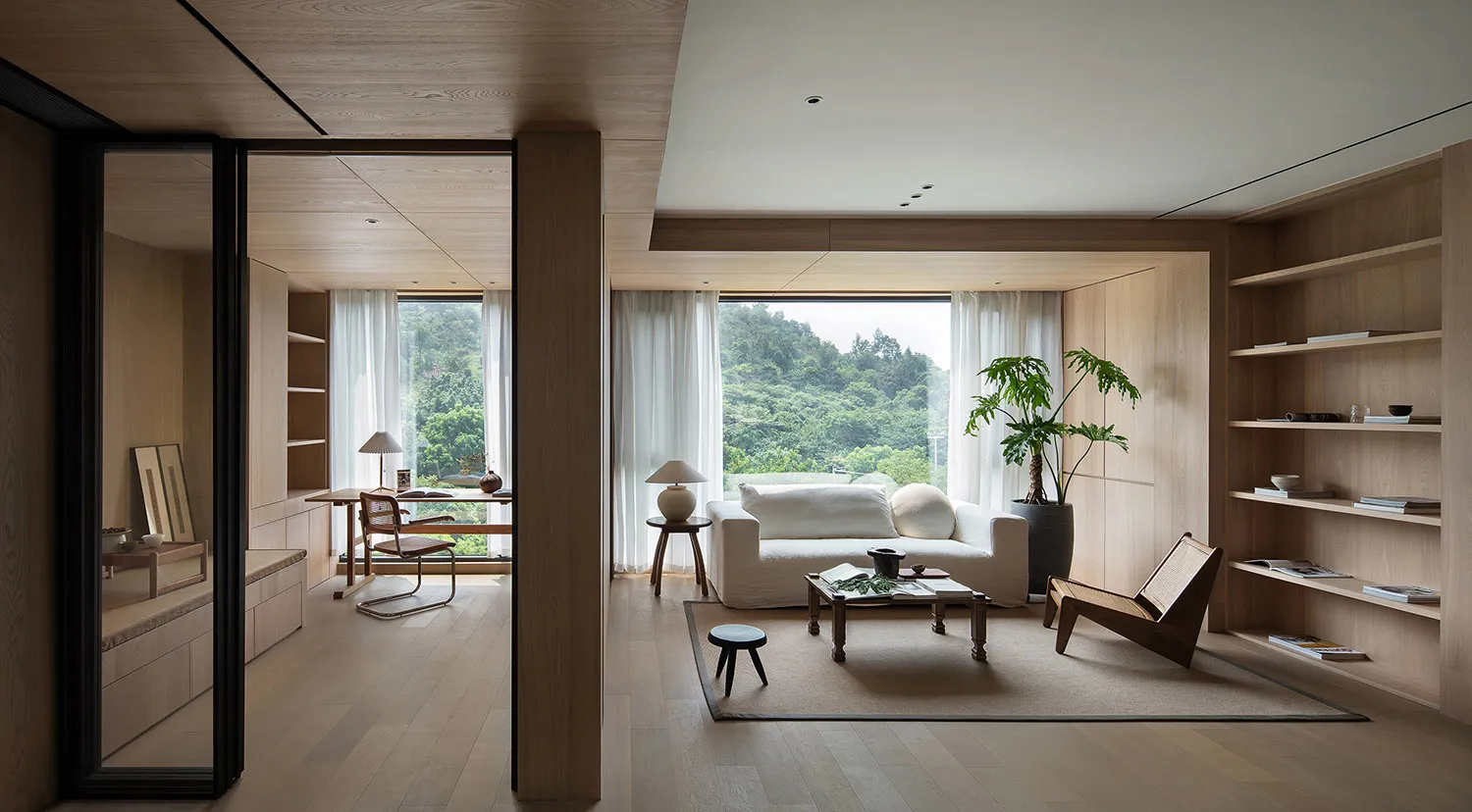 A modern minimalist living room featuring a white couch, wooden accents, and glass windows overlooking a lush green landscape