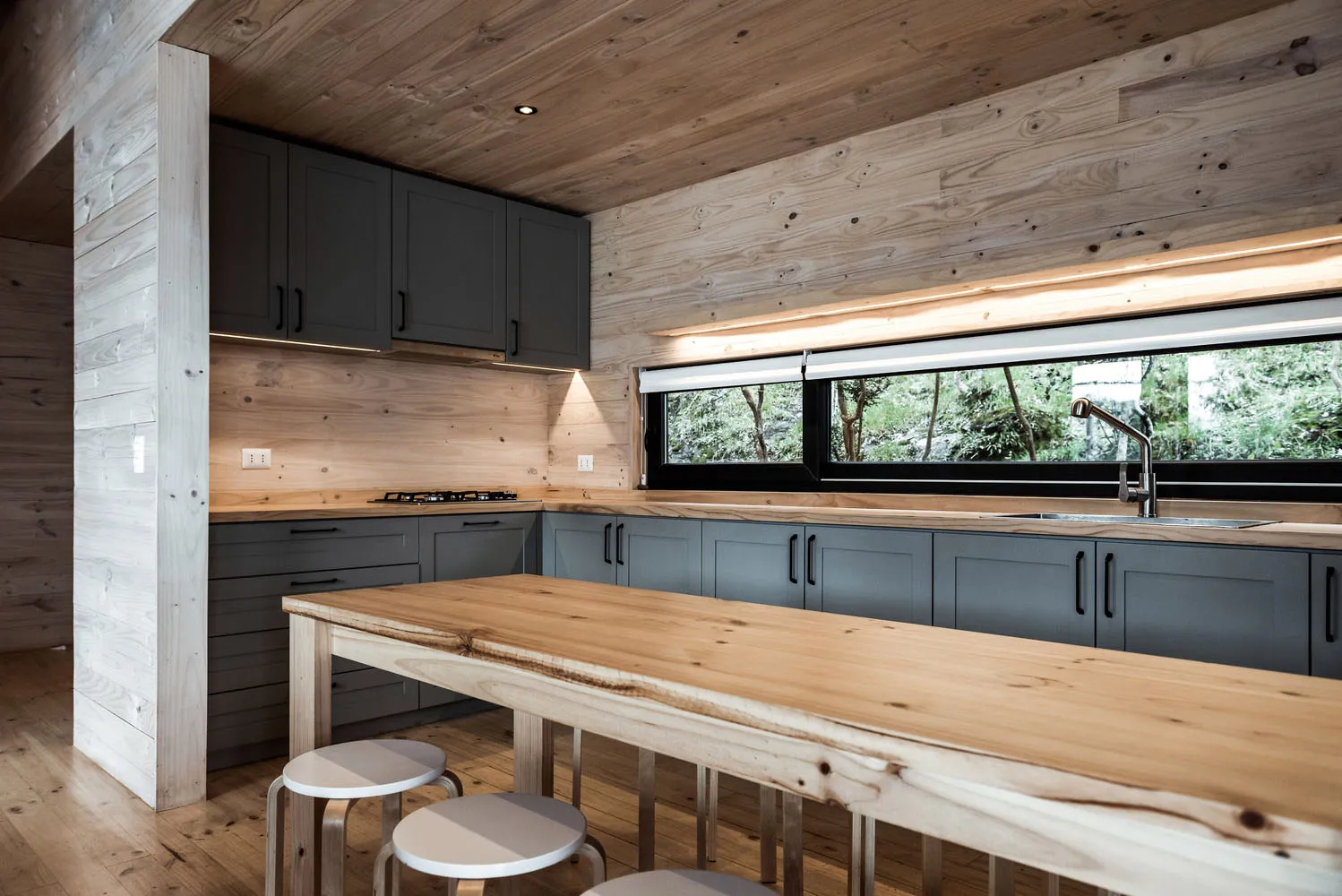 A minimalist kitchen featuring wooden cabinets and countertops, with a long wooden table in the foreground and large windows