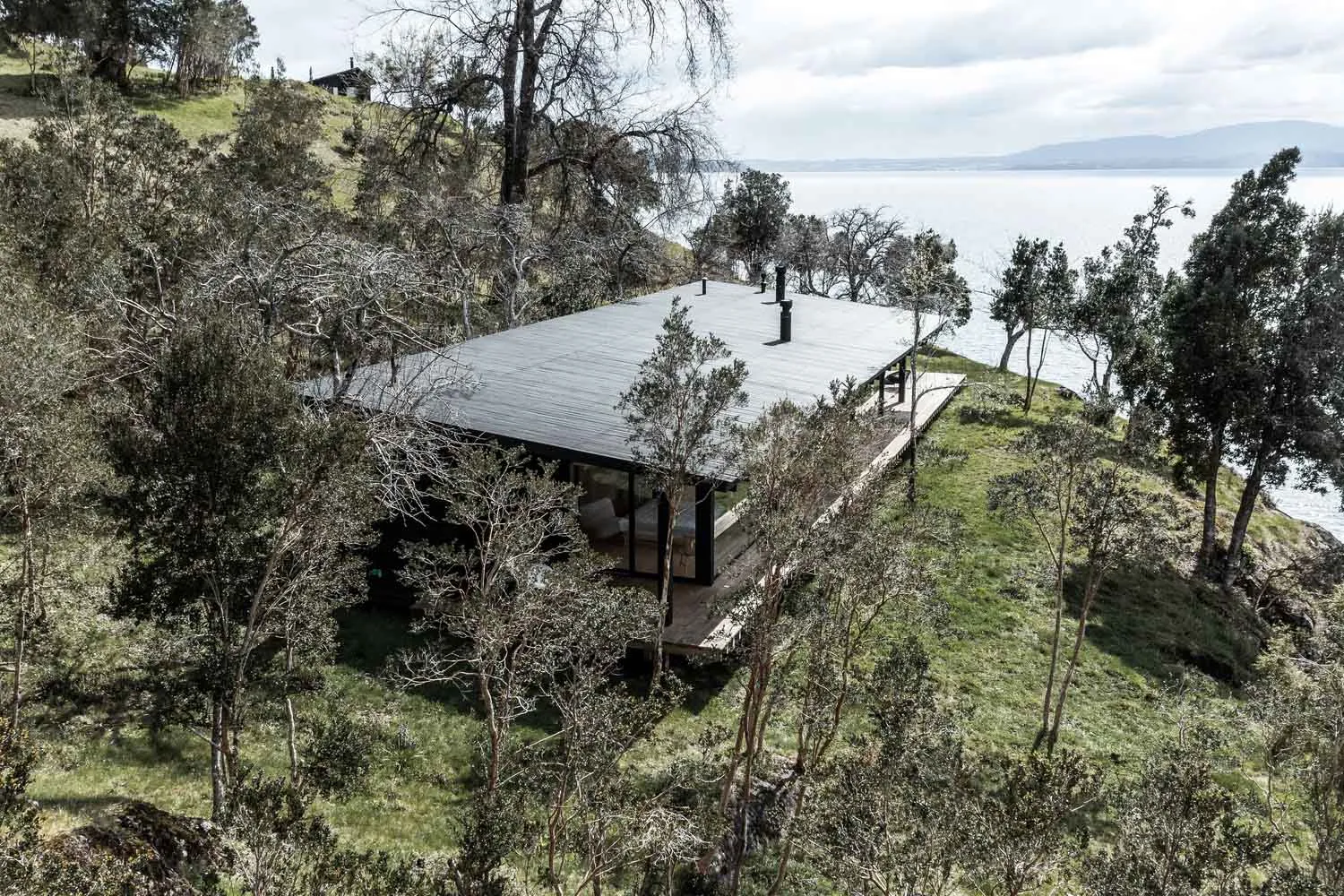 A modern wooden-roof house overlooking water, surrounded by greenery and trees.