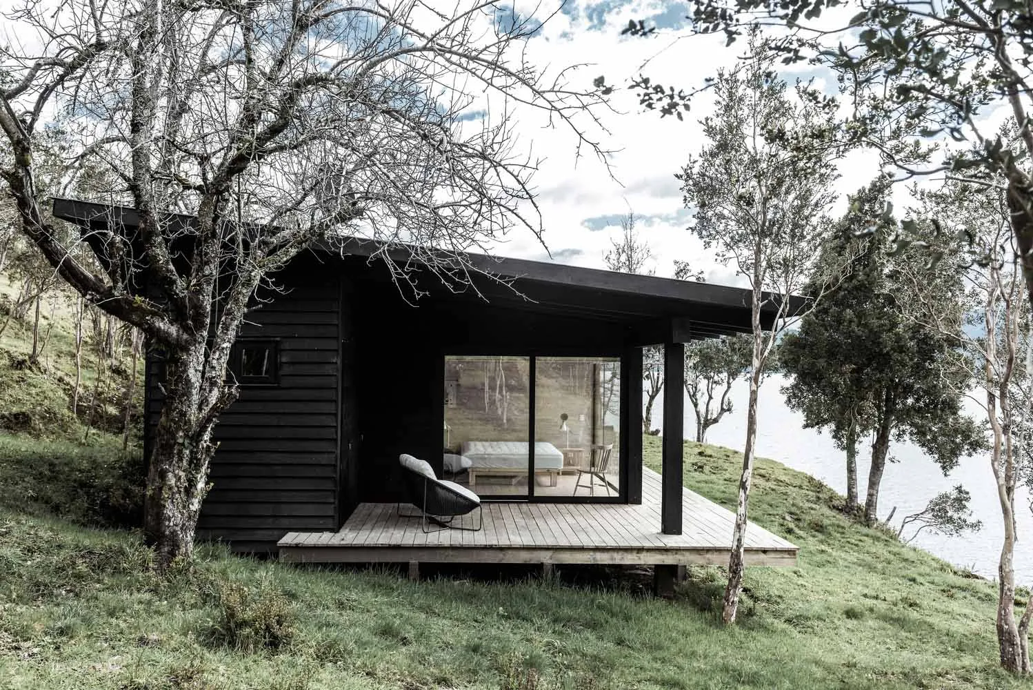 A minimalist black cabin with a wooden deck and glass windows, situated by a body of water.