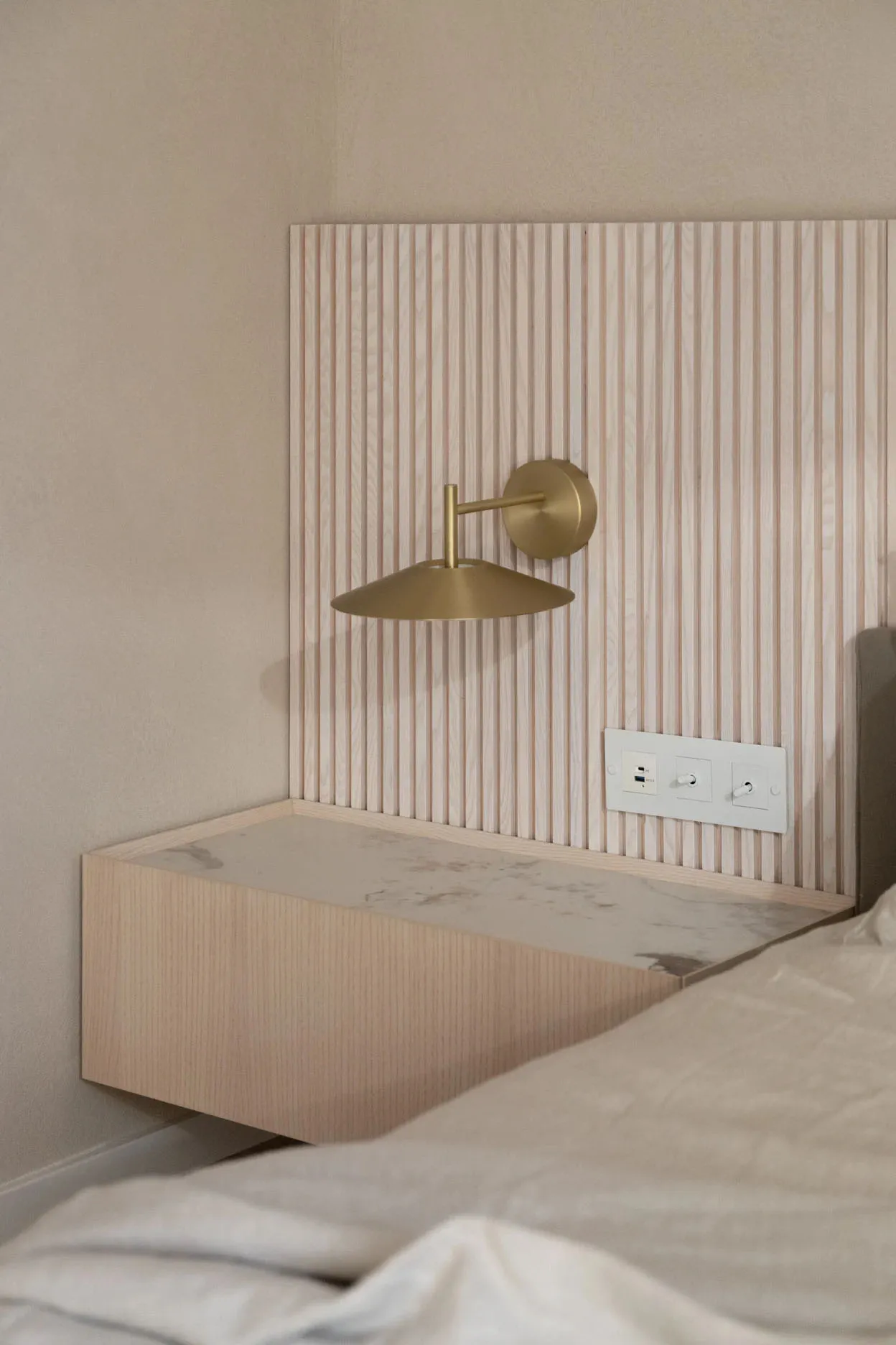 Modern bedroom featuring minimalist design with brass wall lamp, marble surface, and wooden paneling.