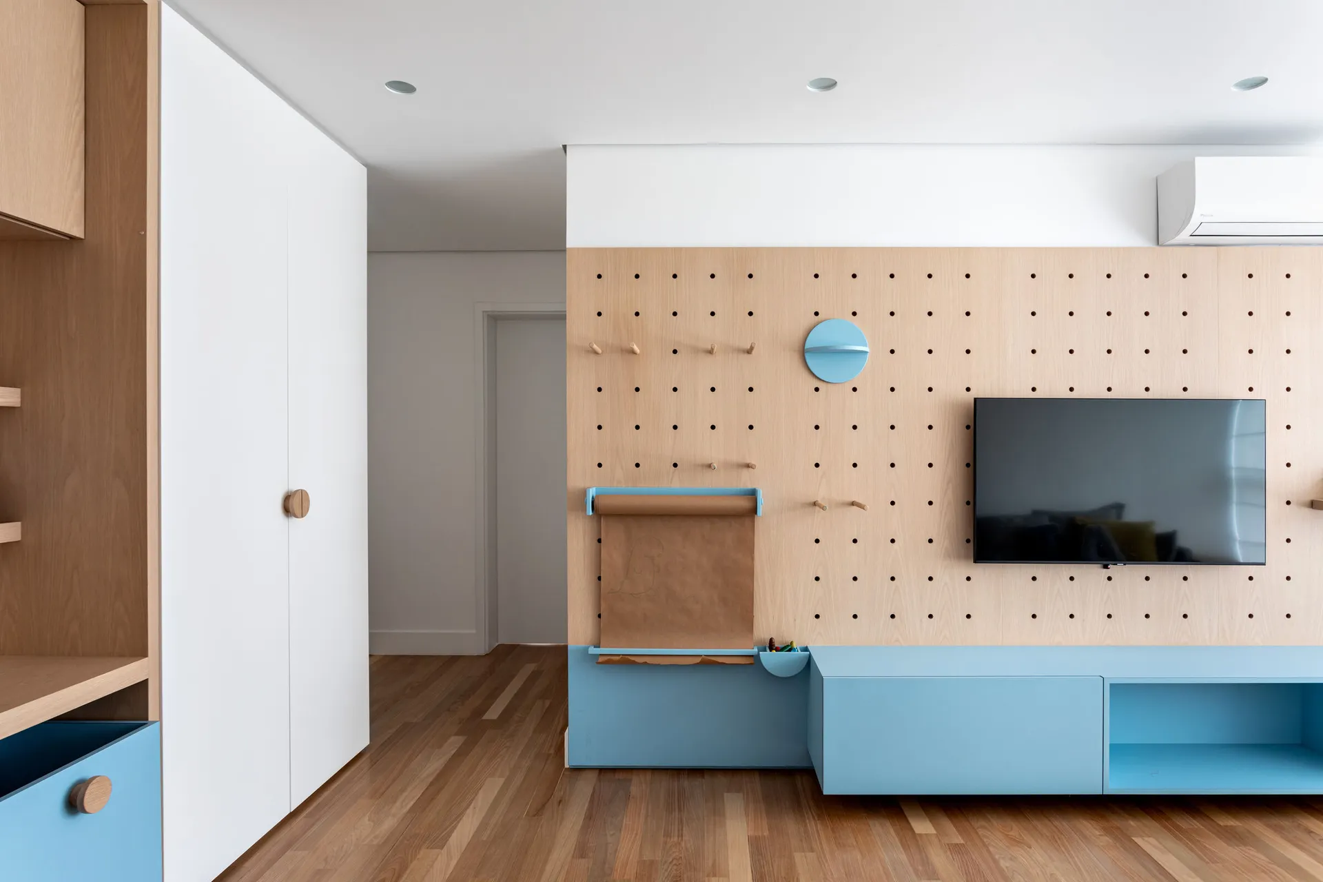 A minimalist living room with blue and wood accents featuring a pegboard wall, a blue cabinet, and a mounted television.