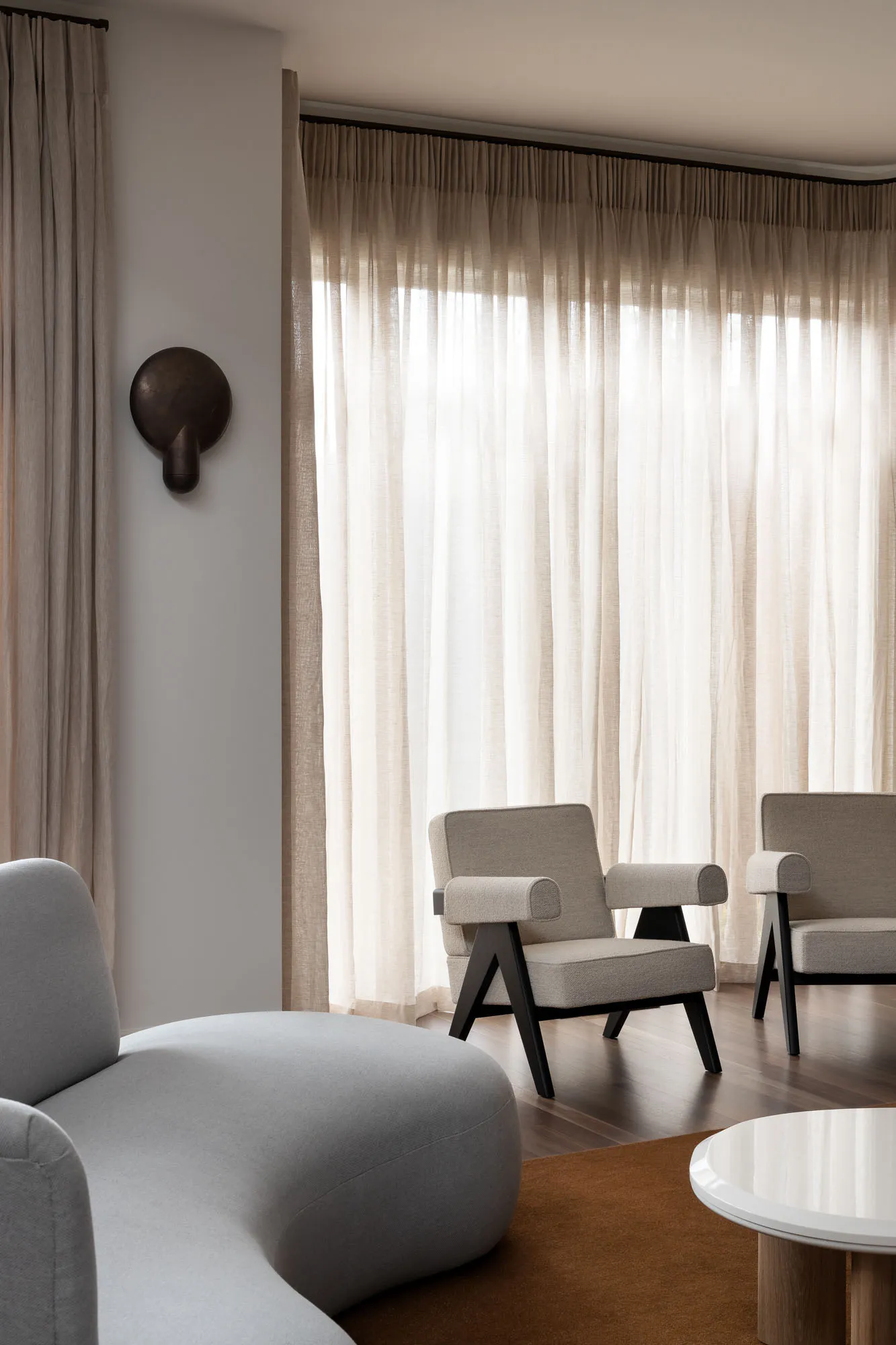 A minimalist living room featuring fabric curtains, a gray sofa, and two modern armchairs with black legs.