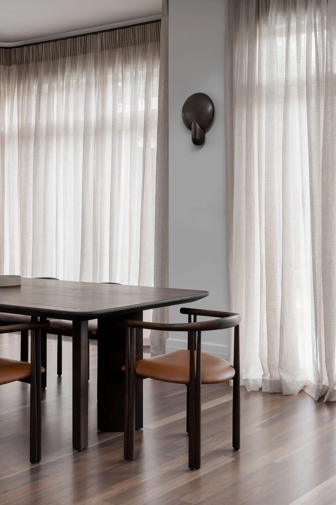 A modern dining room featuring wooden furniture, leather seating, light-colored sheer curtains, and wall-mounted light fixtur