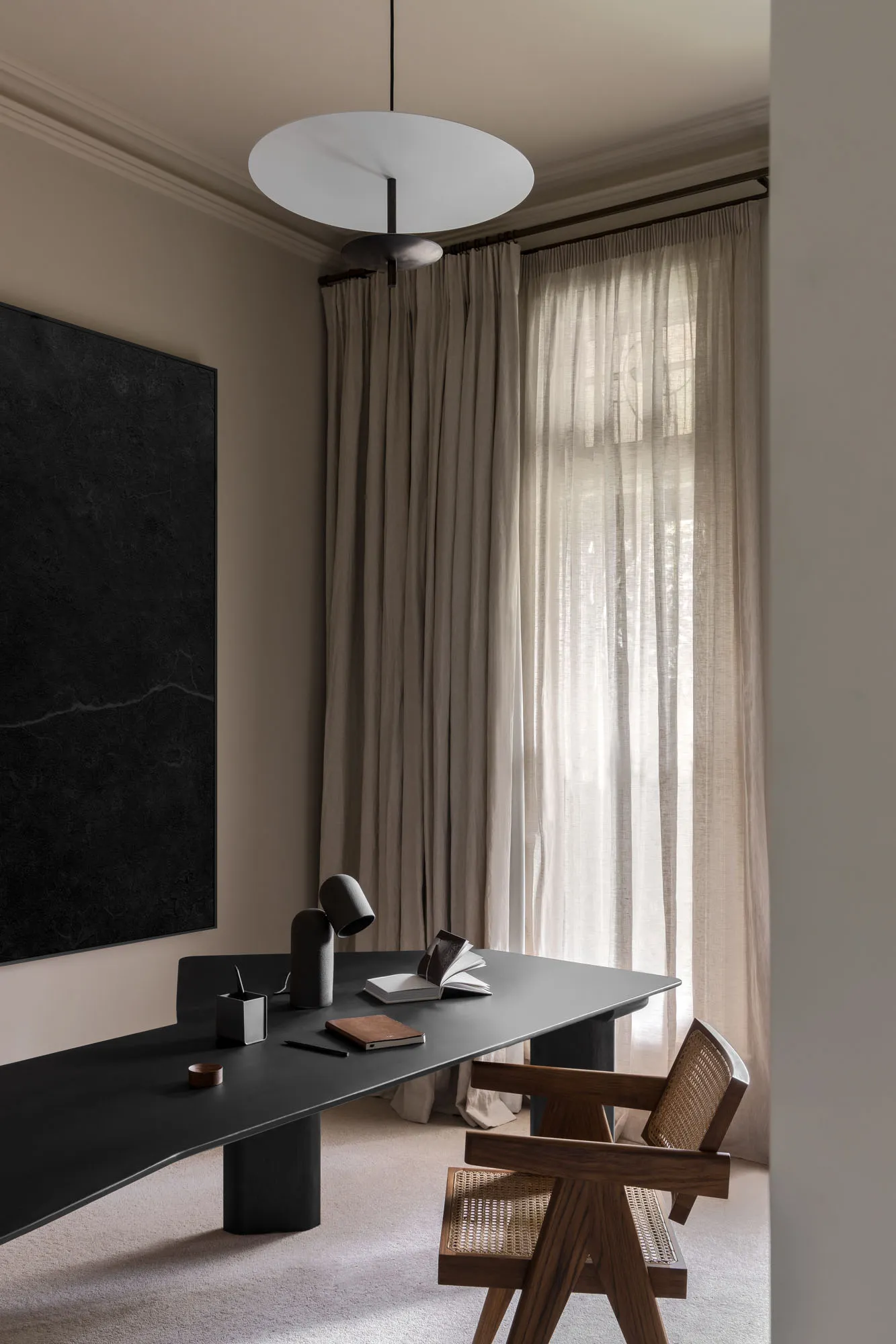 A modern home office featuring a long black desk, a wooden chair, and a linen curtain by the window.