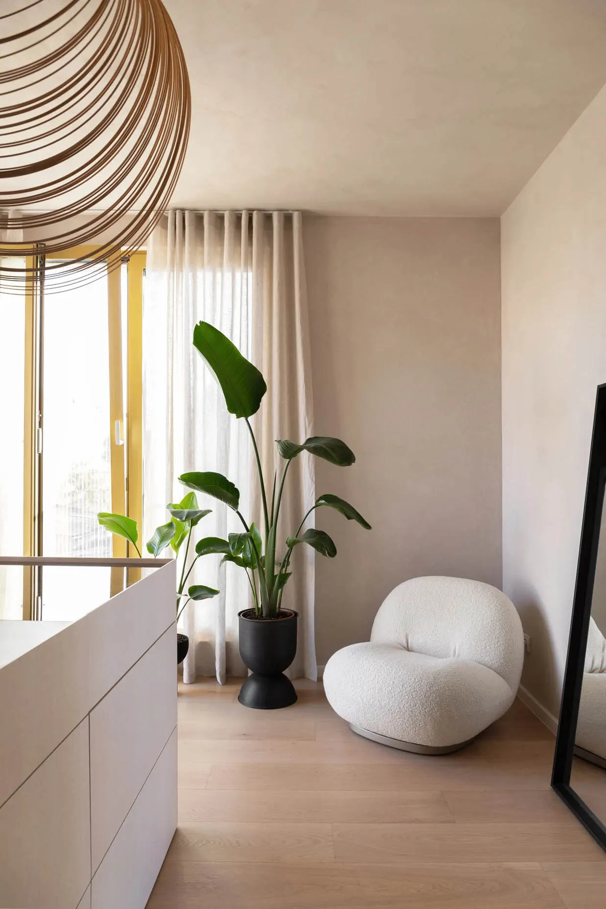 A modern living space with light-colored flooring, minimalist furnishings, and a large window with sheer curtains.