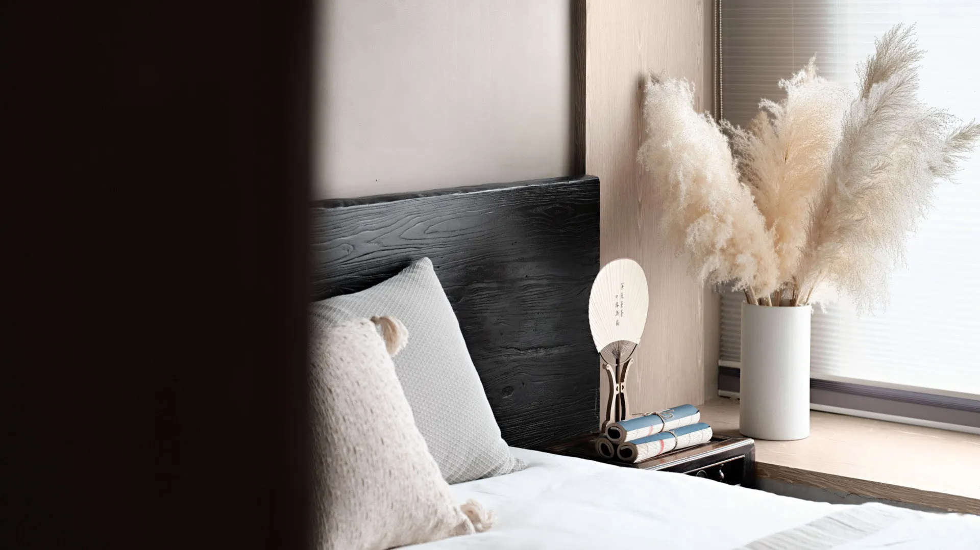 A minimalist bedroom with a black wood headboard, white bedding, and dried pampas grass in a vase.