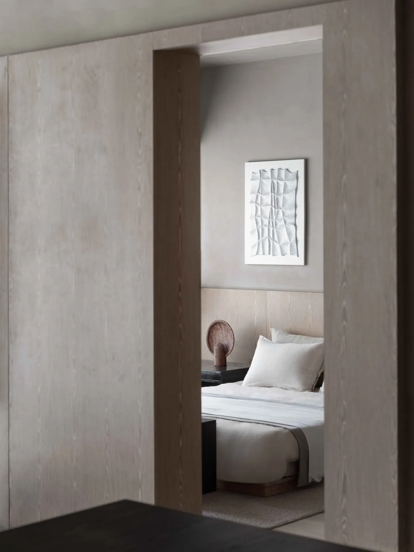 A minimalist bedroom with a wooden headboard and grey walls, featuring a white bedspread and decorative vase on the nightstan