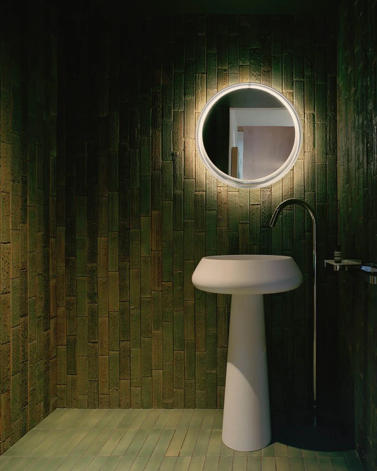 A minimalist bathroom with a green-tiled wall and white freestanding sink.
