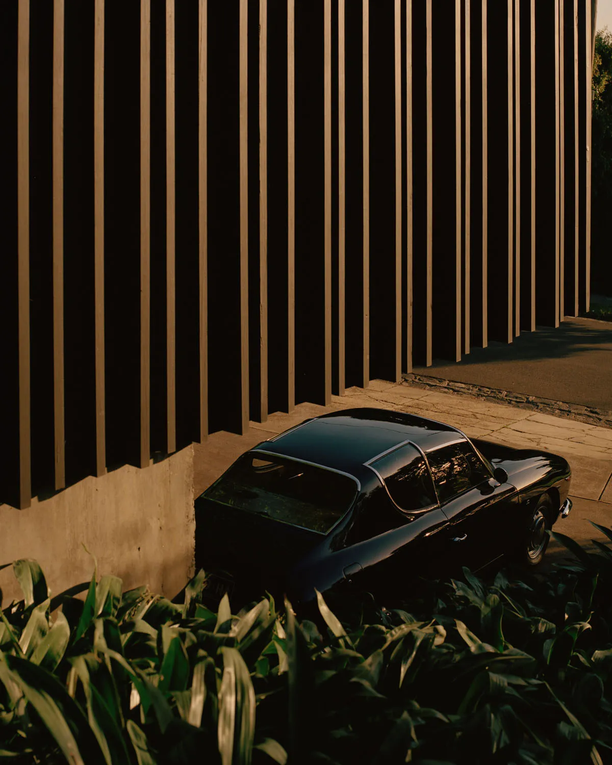 Black vintage car parked next to a modern metal building, surrounded by greenery.
