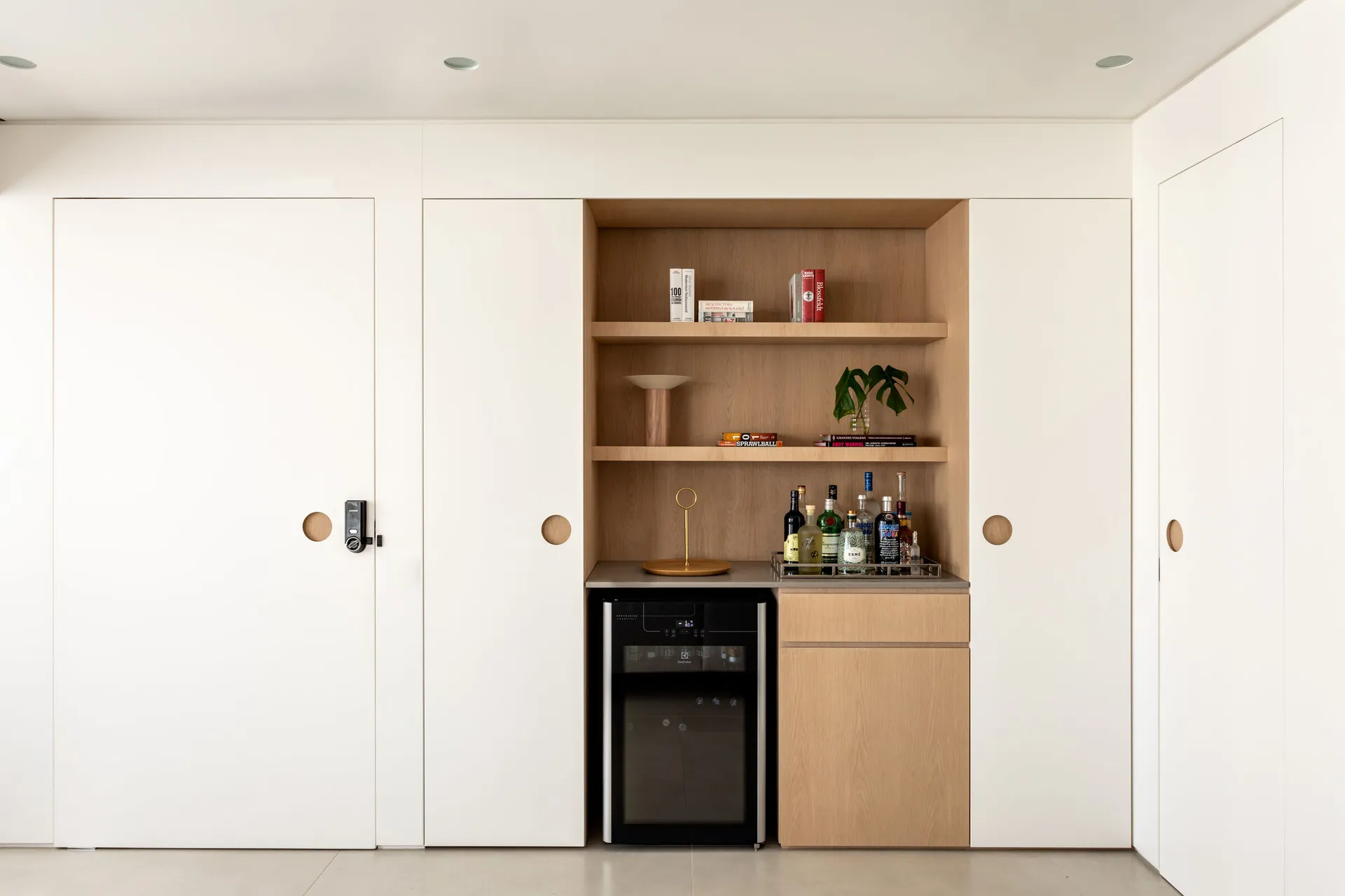 A modern, minimalist home bar area with wooden shelves, a mini fridge, and bottles of alcohol.