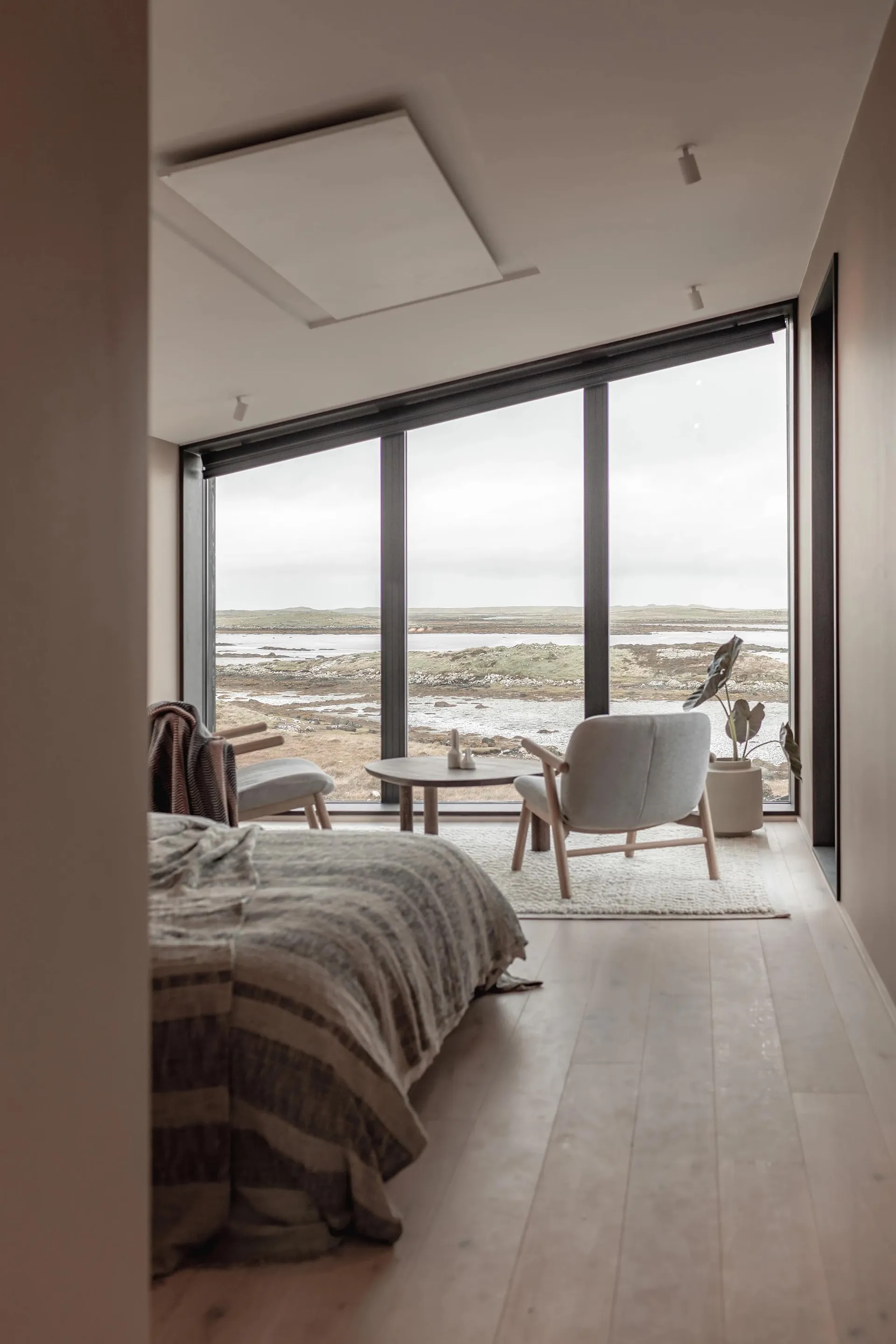 Minimalist bedroom with white walls, wooden floor, large window overlooking a scenic landscape, and simple furniture.