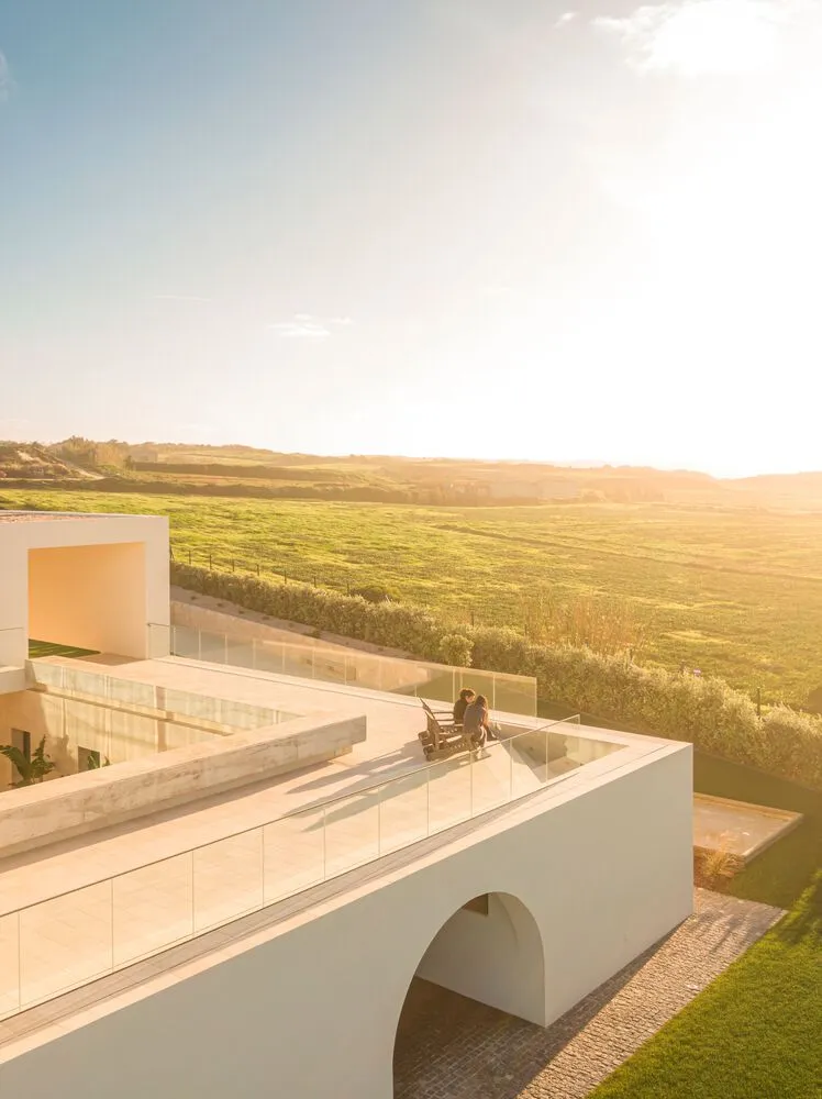 A modern residential veranda with concrete flooring and glass railings overlooking green fields at sunset.