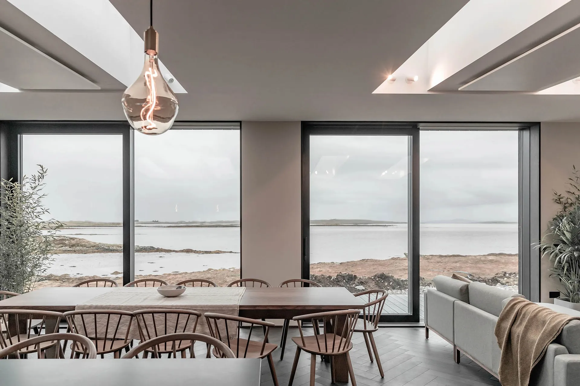 A modern residential dining room featuring a long dining table with wooden chairs, large glass windows with a coastal view, a