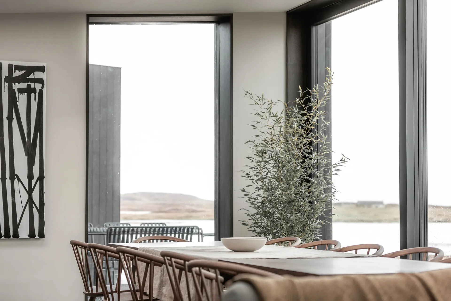 A minimalist dining room with wood furnishings and a large window with greenery outside.