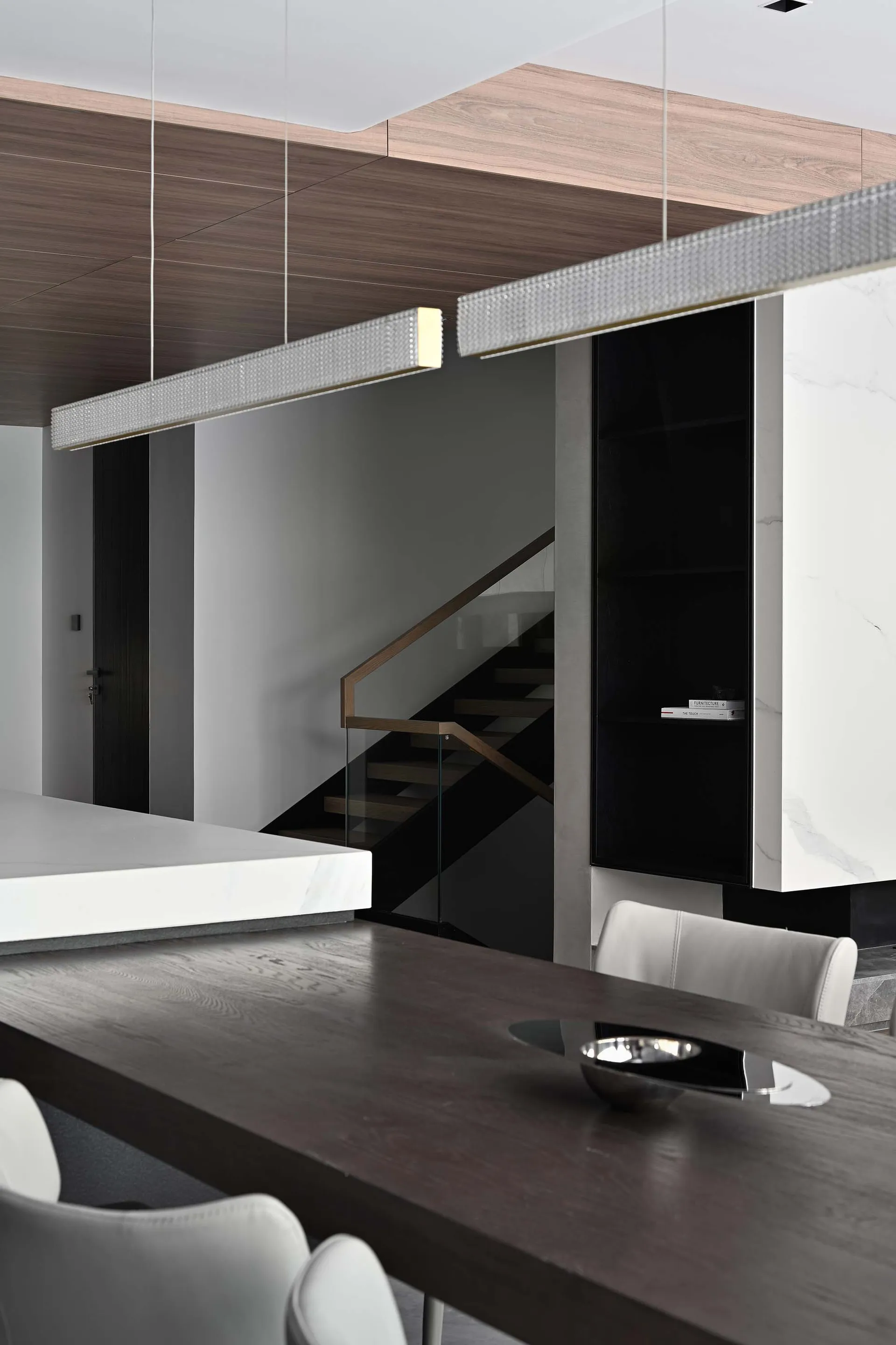 Modern minimalist dining room featuring metallic lighting above a wooden table with white chairs and a glass staircase in the