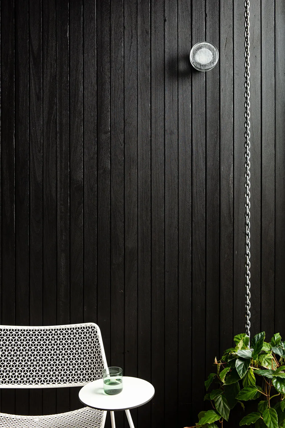 A minimalist white metal chair with black wooden paneling and green plants in the foreground.