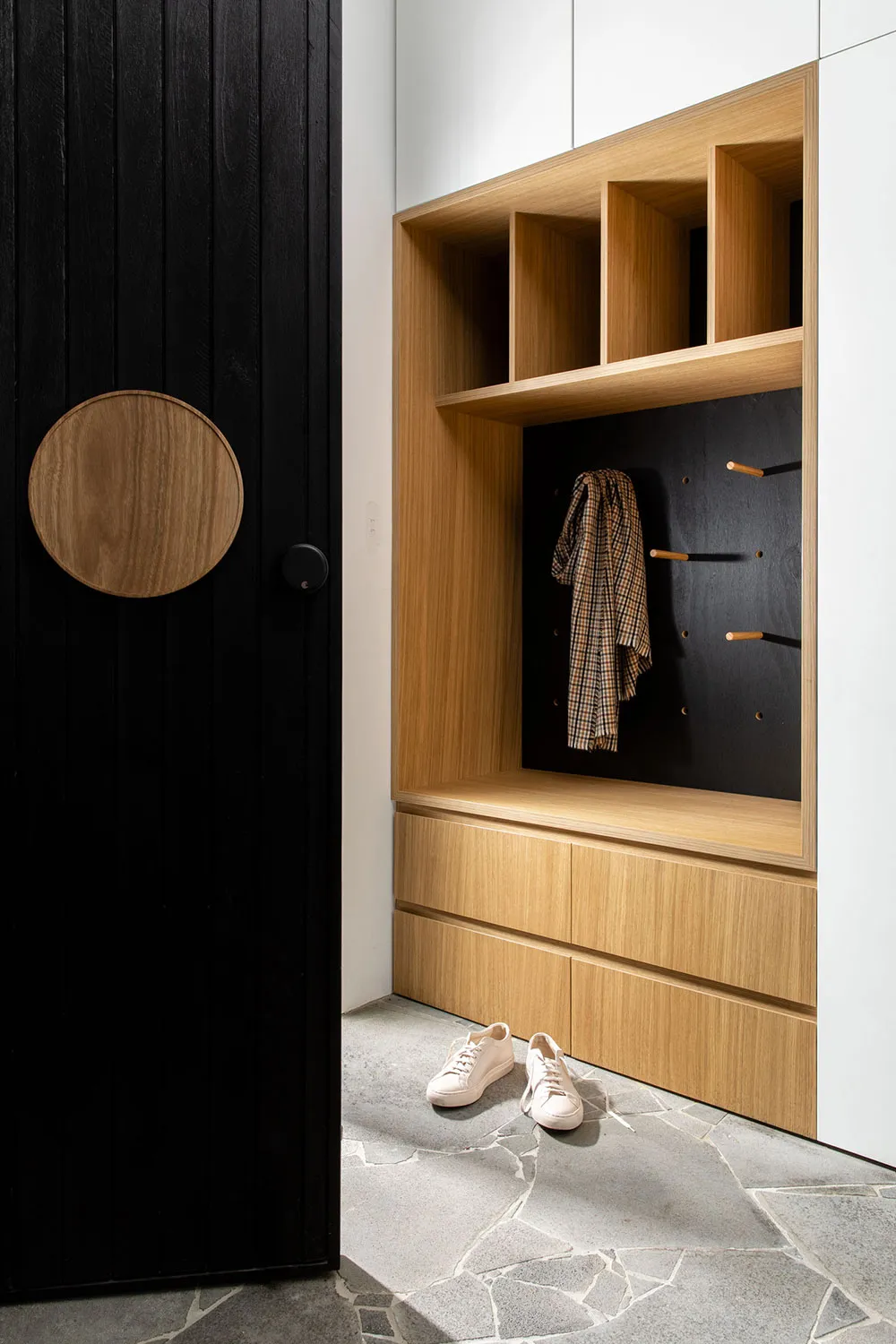 A modern entryway with wooden storage shelves, a black door with a circular wooden element, and shoes on the floor.