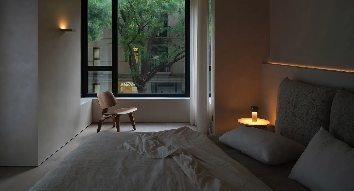 A minimally decorated bedroom featuring a textured fabric bedspread, a wooden chair, and a small table lamp with an orange li
