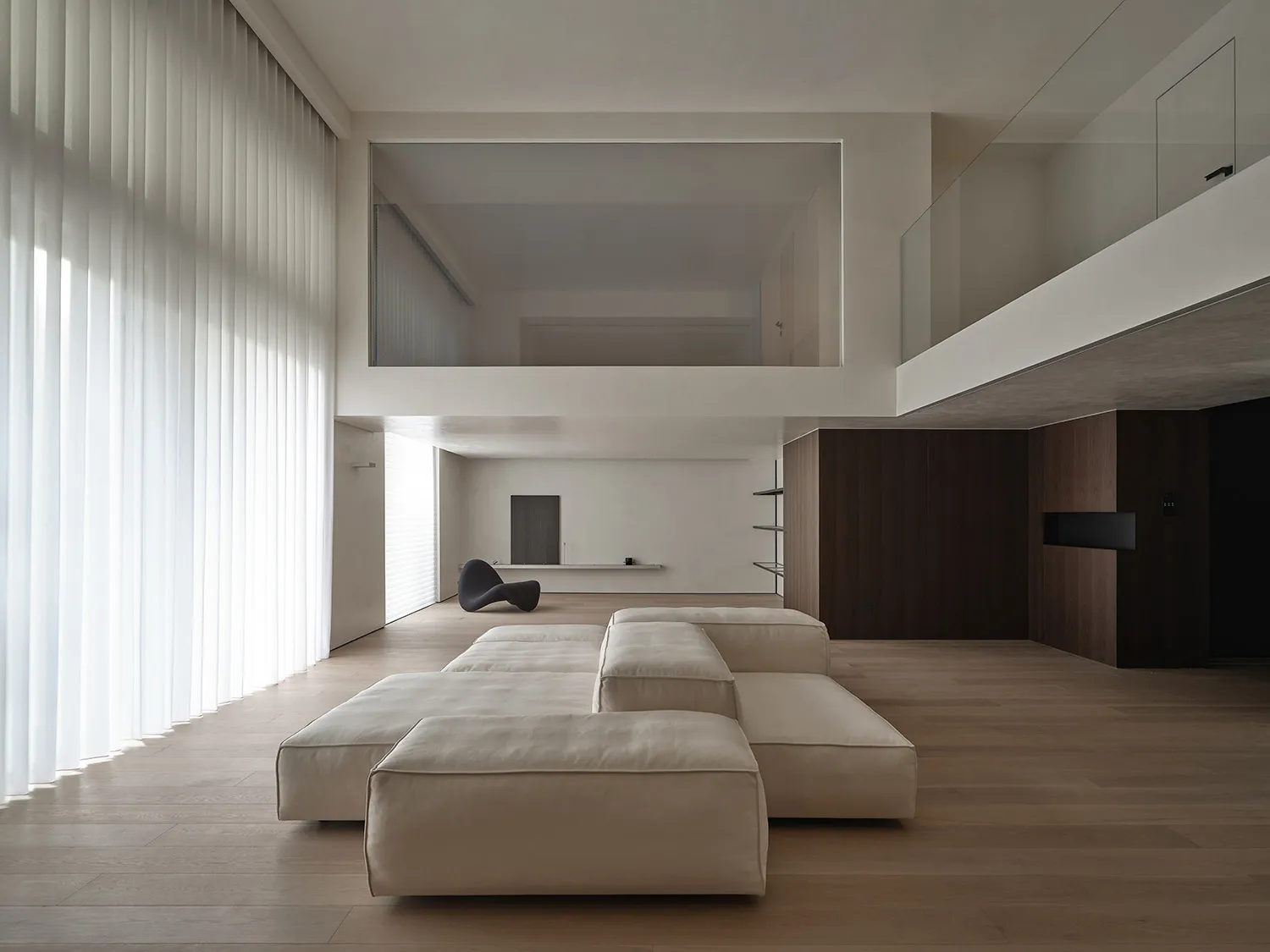 A modern minimalist living room with large windows and a wooden floor, featuring a beige modular sofa and a glass partition.