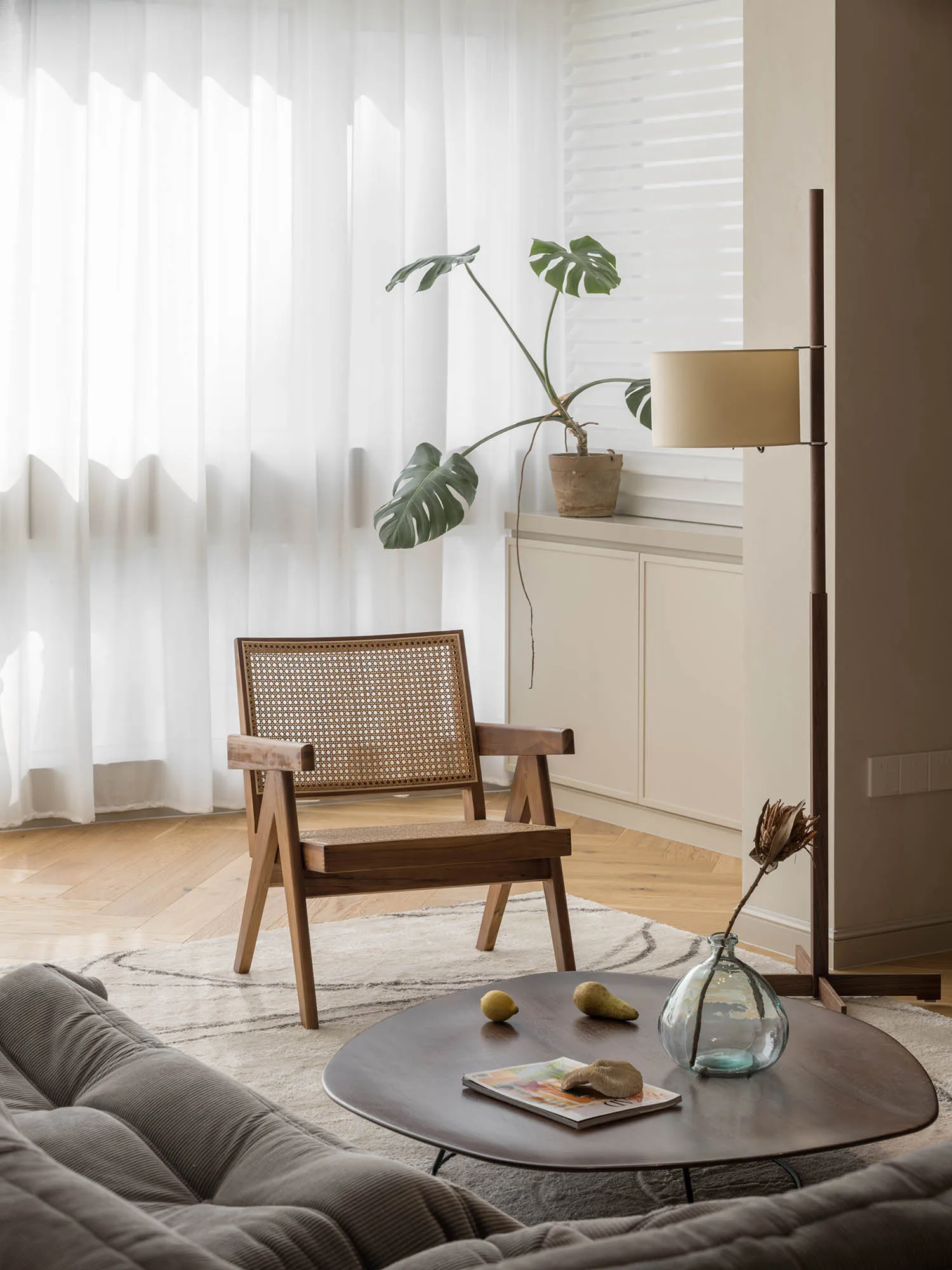 A modern minimalist living room featuring a wooden armchair, a round wooden table with fruits and a magazine, and a floor lam