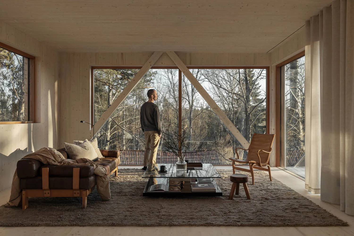 A minimalist living room with wooden beams and large windows showcasing a forest view.