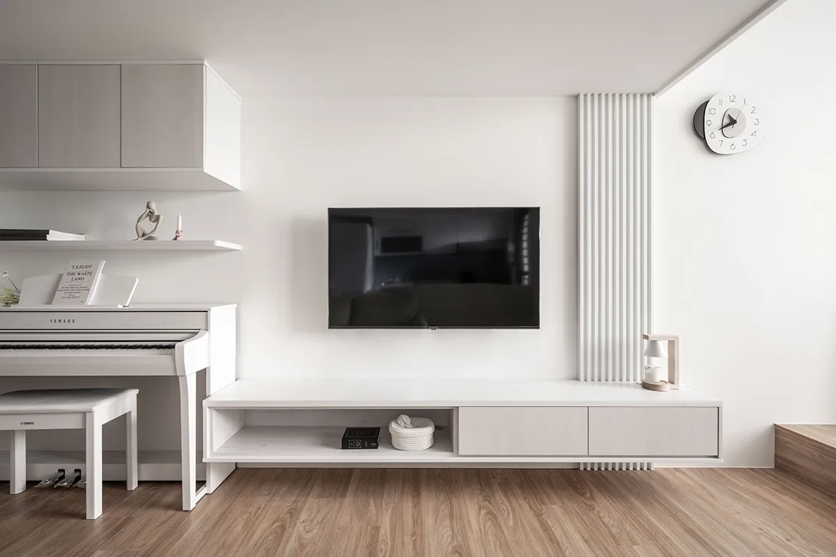 Modern living room with white walls, furniture, and floor, featuring a white piano, a TV mounted on the wall, and a white clo