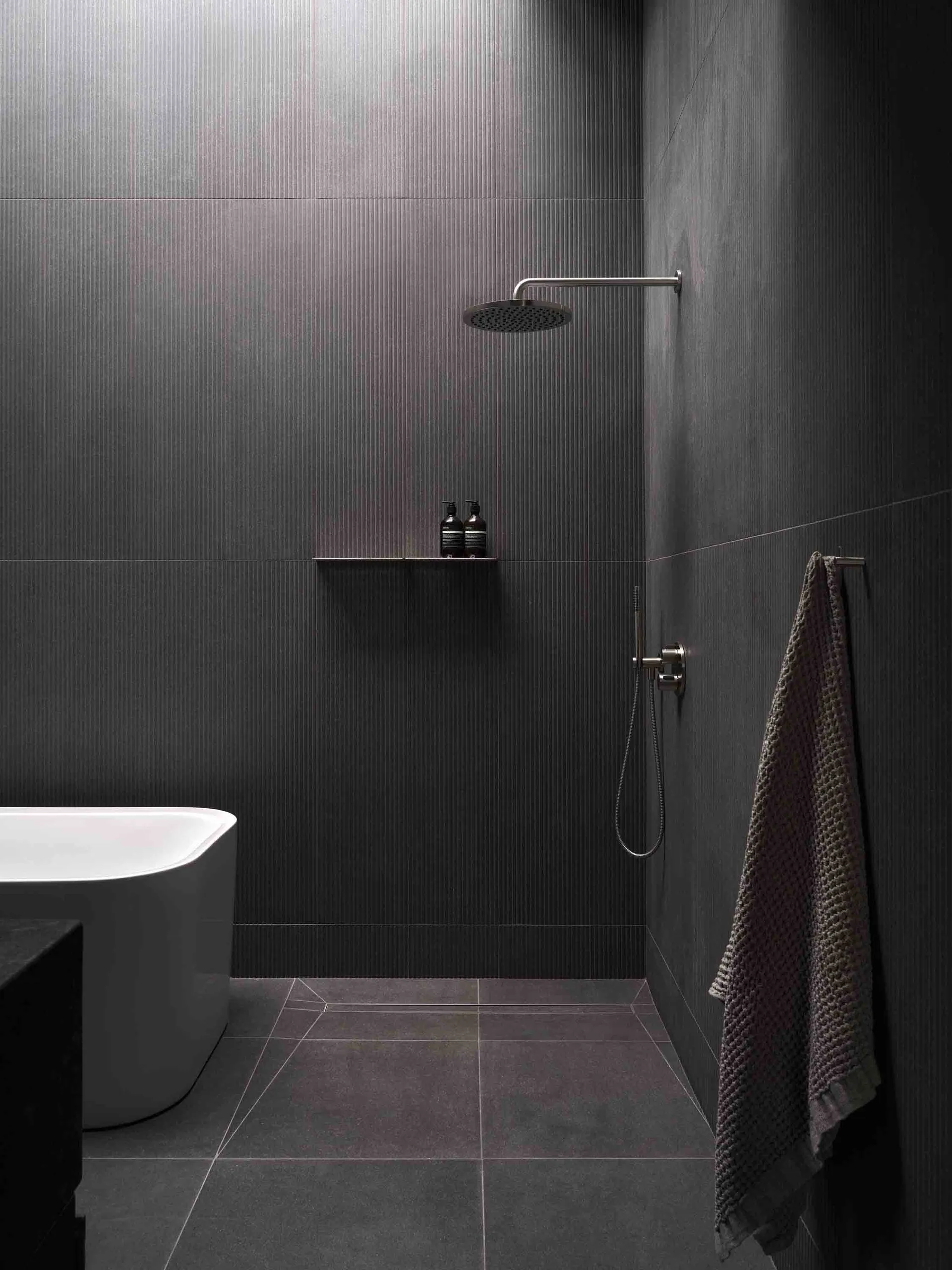Modern minimalist bathroom with dark tiles and concrete floor, featuring a freestanding tub and walk-in shower.