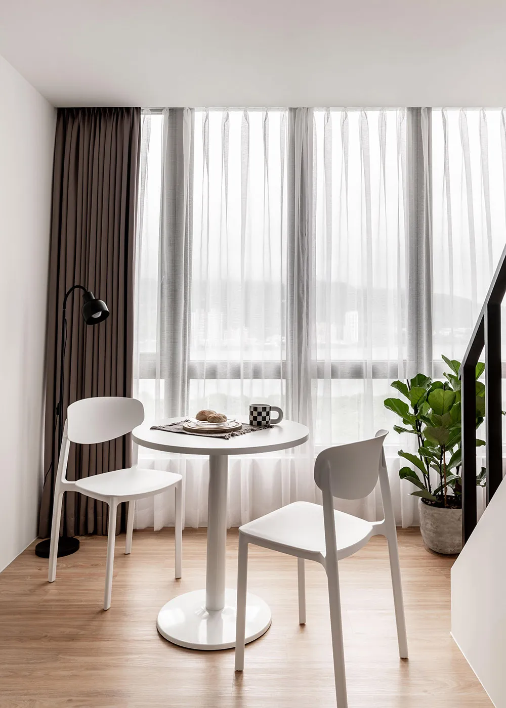 A minimalist dining area featuring a round white table with a plate, mug, and bread, surrounded by two white chairs, under a