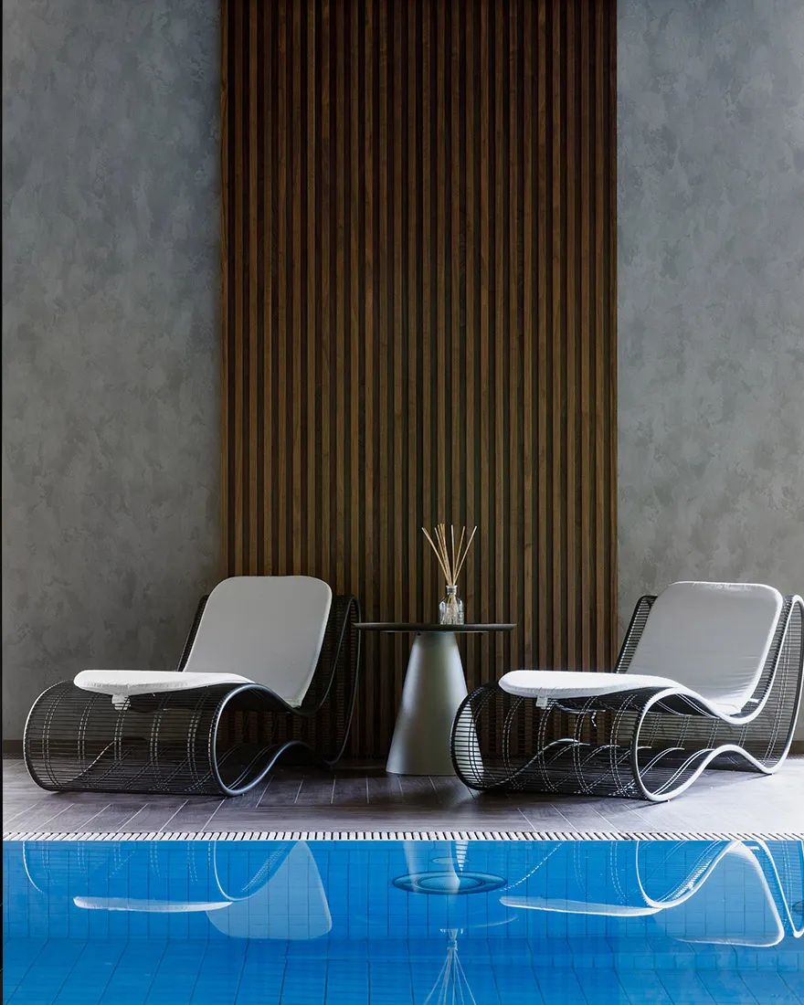 A minimalist poolside seating area featuring modern metal chairs and a table with a vase of reeds, placed adjacent to a swimm