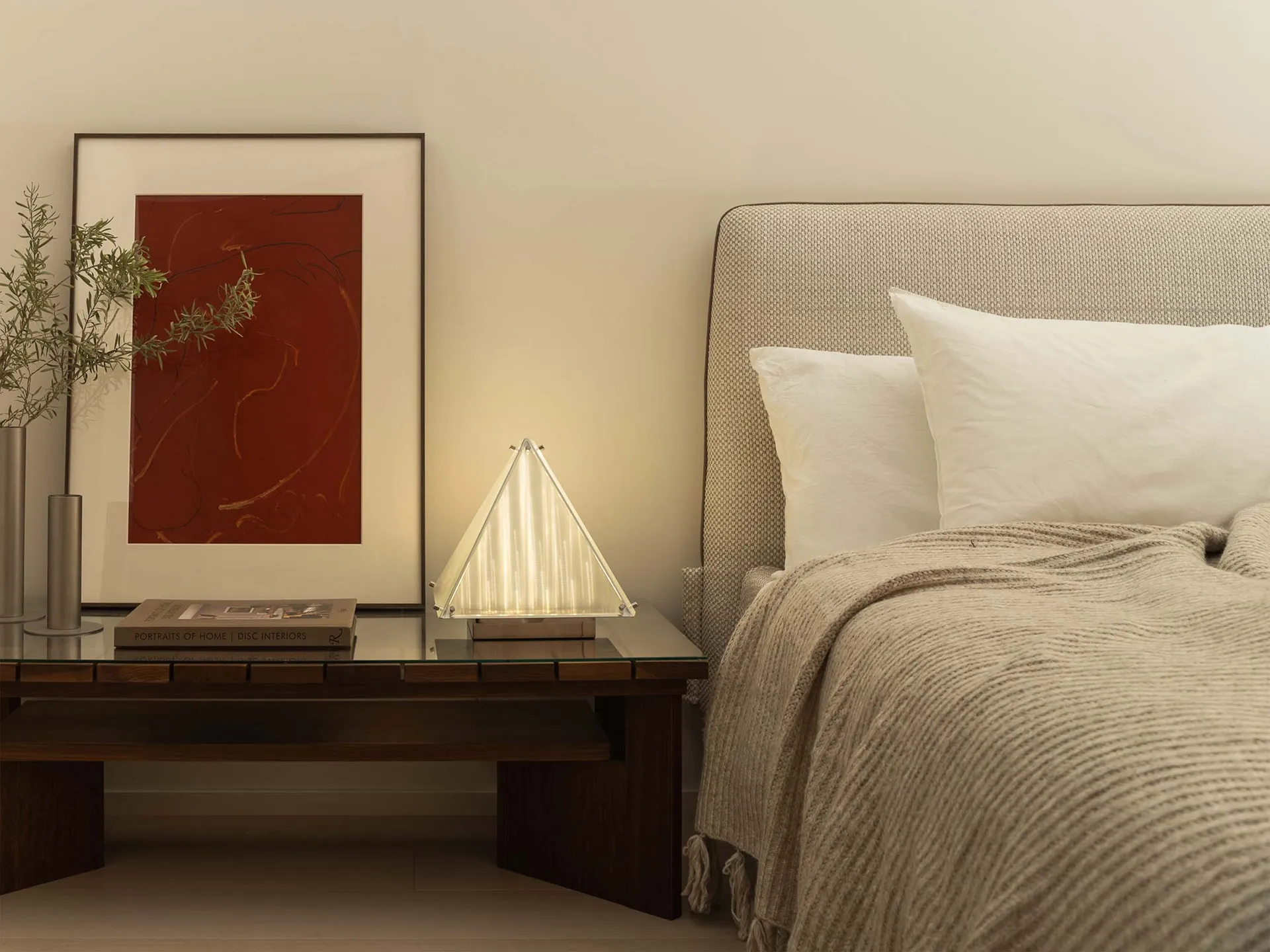 Modern minimalist bedroom featuring soft textiles on the bed, with a soft lighting lamp on the bedside table and a framed abs